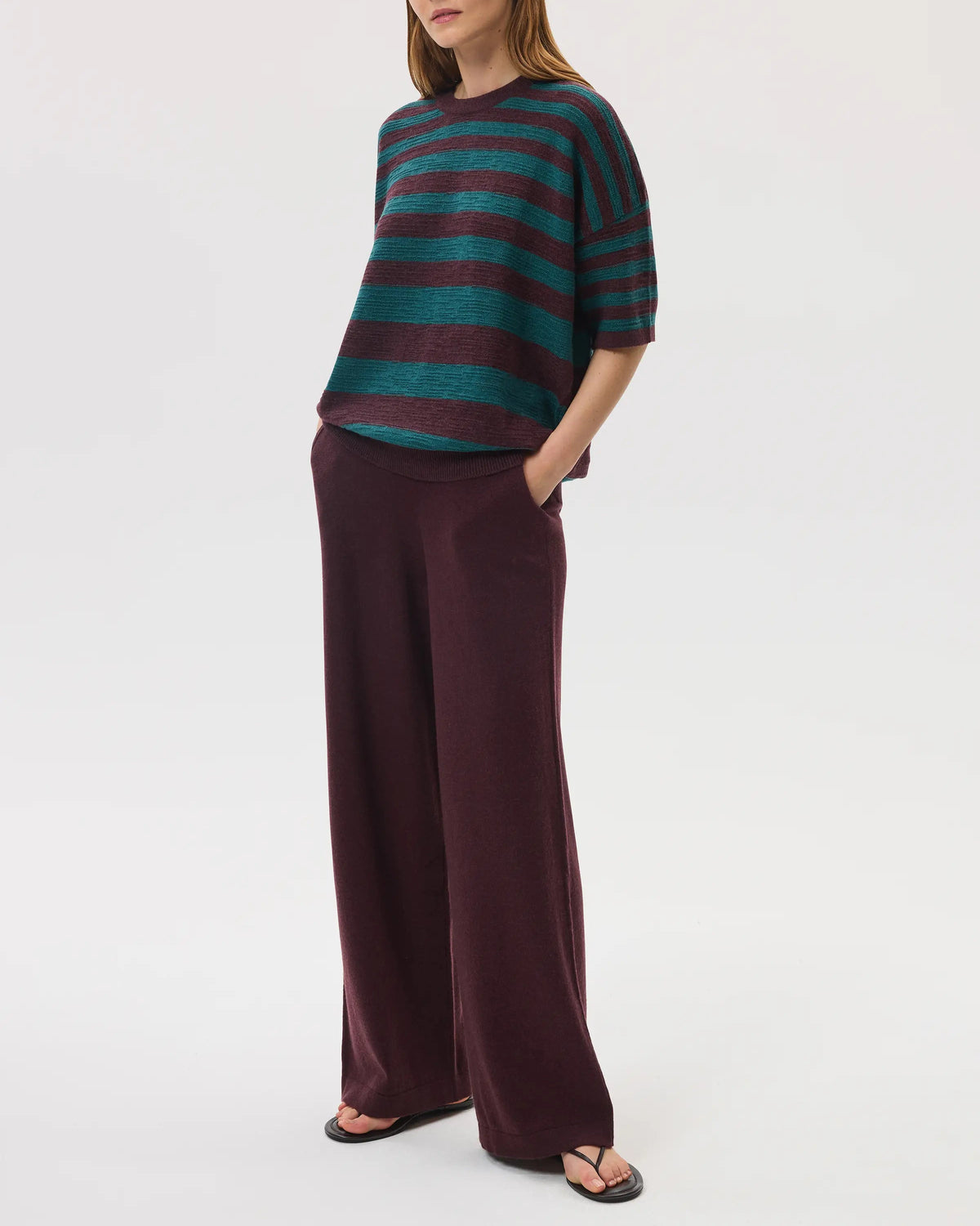 Female model wearing Johnstons of Elgin SS25 Unisex Knitwear Rosewood & Emerald Striped Oversized Cashmere T-Shirt with rosewood wide leg trouser on a white background KAA05414Q25125