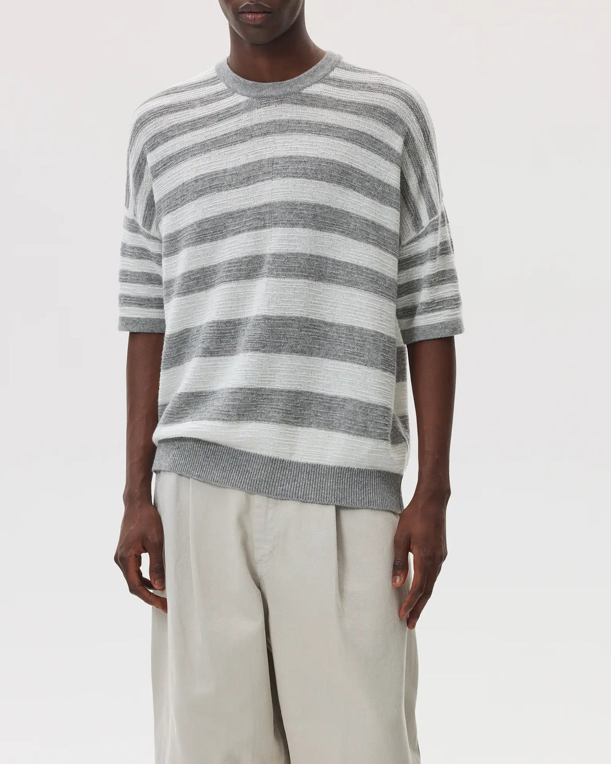 Male model wearing Johnstons of Elgin SS25 Unisex Knitwear Light Grey & White Textured Stripe Oversized Cashmere T-Shirt with cream trousers and sandals on a white backdrop KAA05414Q25124