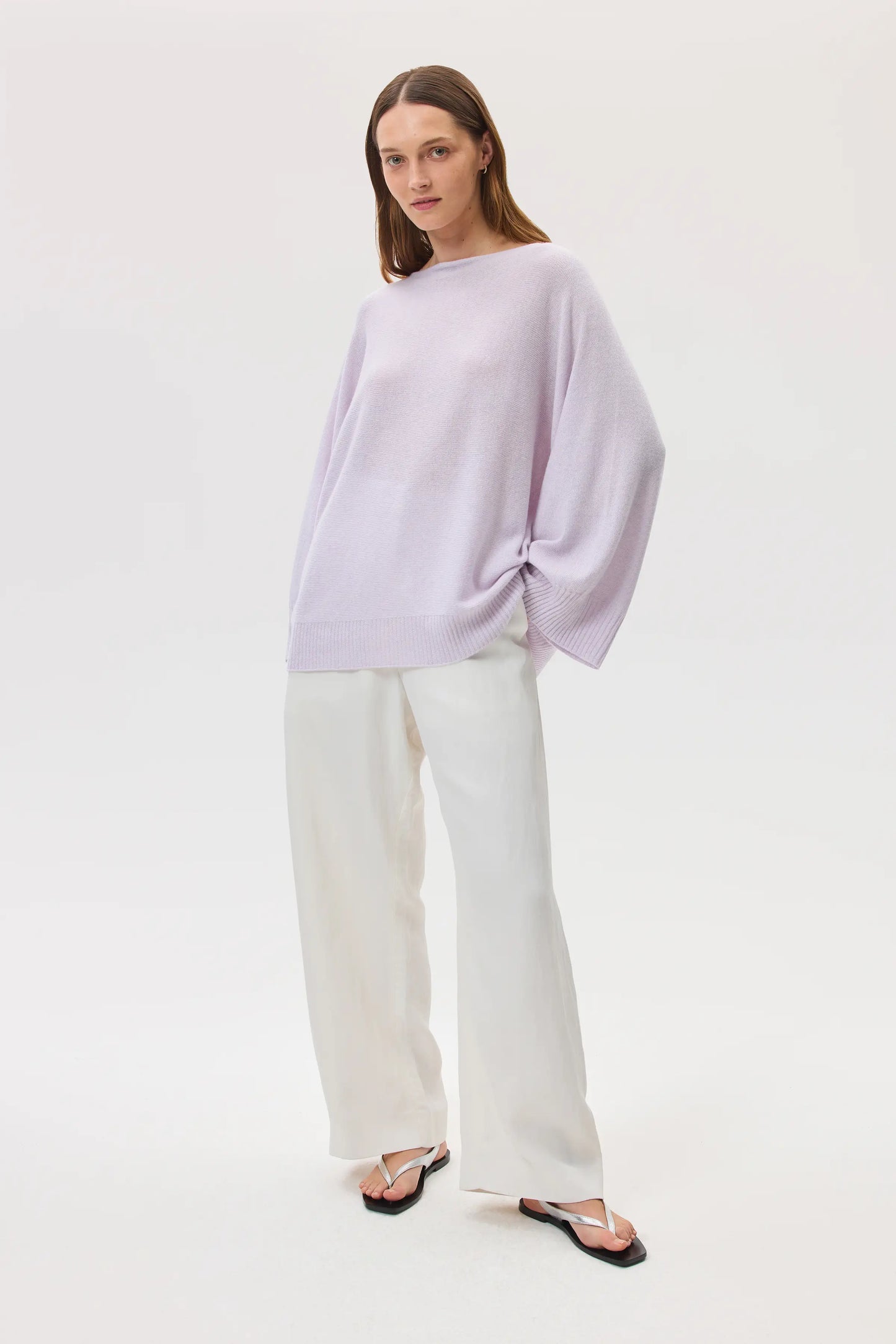 Model wears Johnstons of Elgin SS25 Women's Knitwear Lavender Cashmere Cape Jumper with white trousers and silver sandals KAI05048HD0086