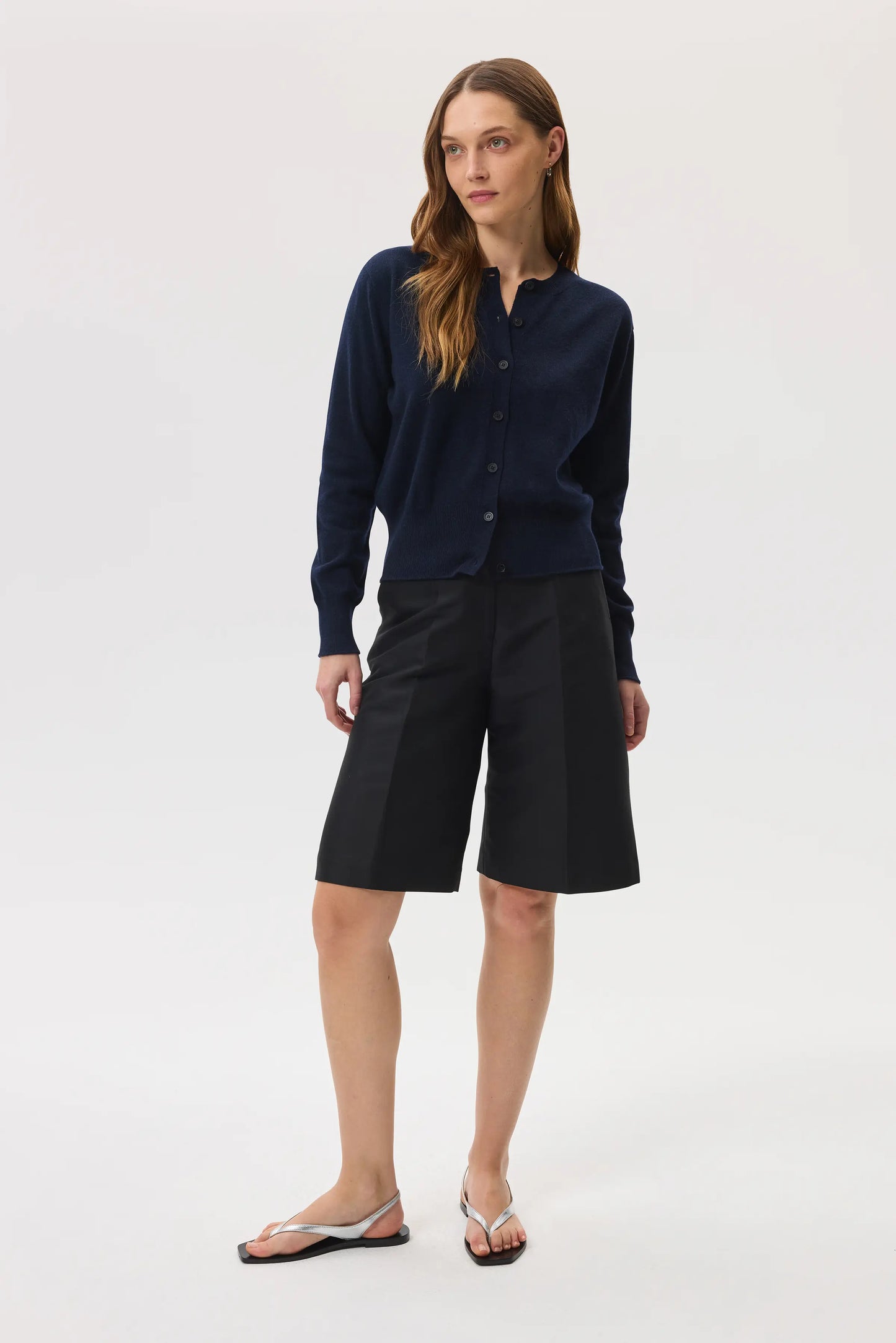 Model wears Johnstons of Elgin SS25 Women's Knitwear Navy Classic Cashmere Cardigan with navy shorts on a white background KAI05274SD7286
