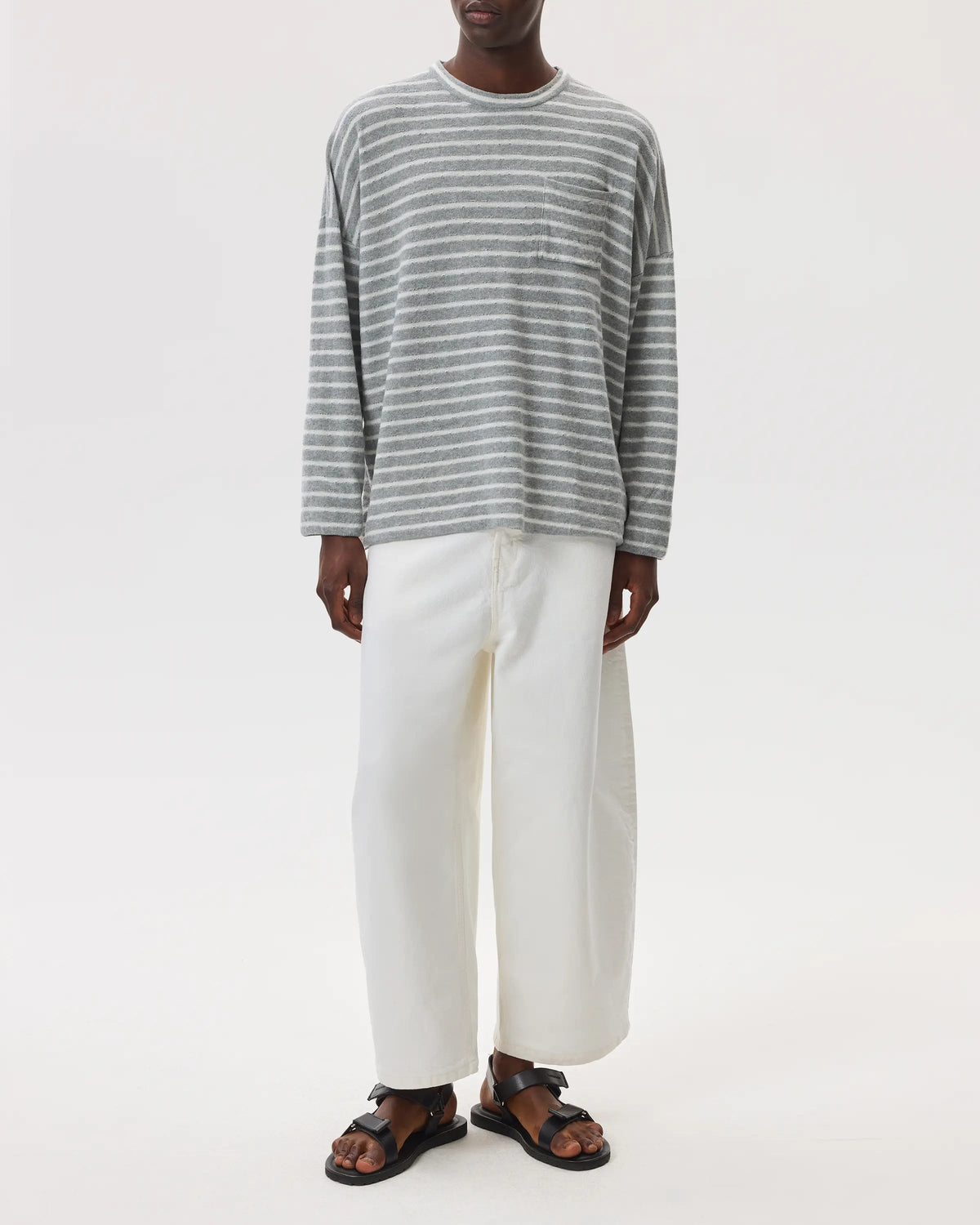 Model wears Johnstons of Elgin SS25 Men's Knitwear Light Grey & White Cashmere Jersey Stripe Oversized T-Shirt with white trousers on a light studio background KAP05442Q25132