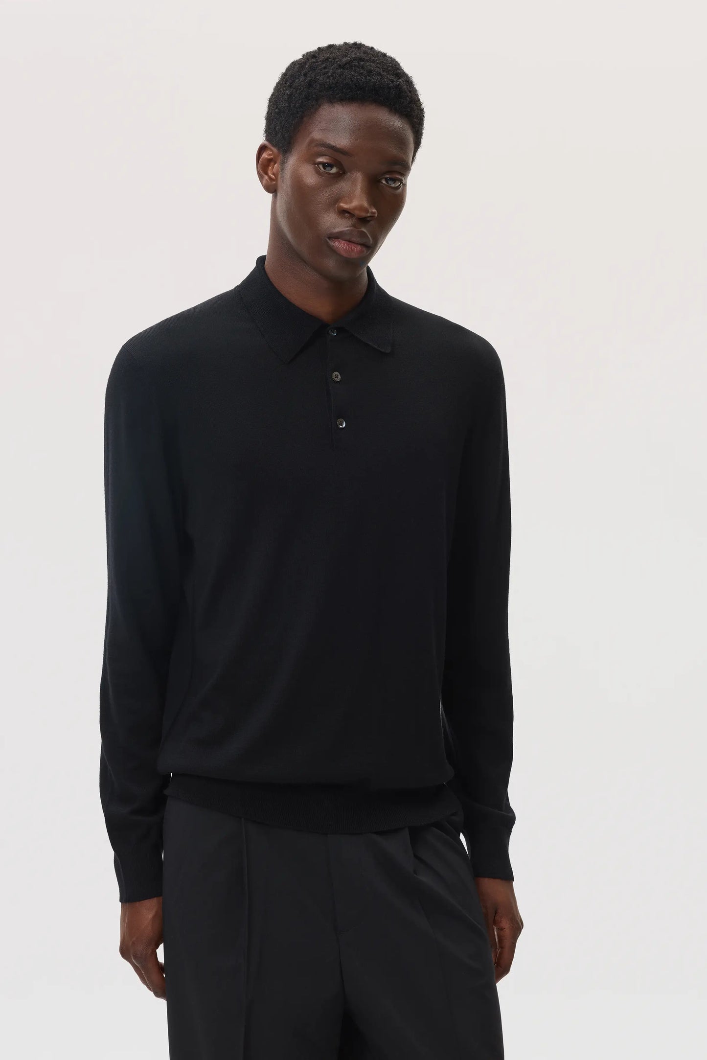 Model wearing Johnstons of Elgin SS25 Men's Knitwear Black Merino Long Sleeve Polo shirt with black trousers
in a white studio KDI00706SA7131