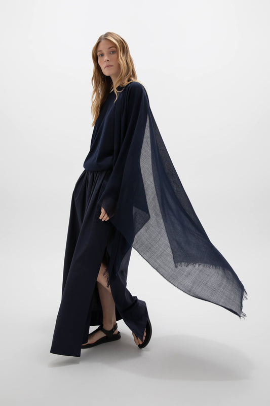 Johnstons of Elgin Lightweight Merino Wool Scarf in Navy worn by a female model over a navy cashmere sweater with a long navy sarong skirt and black sandals against a white background WD001093SD7019