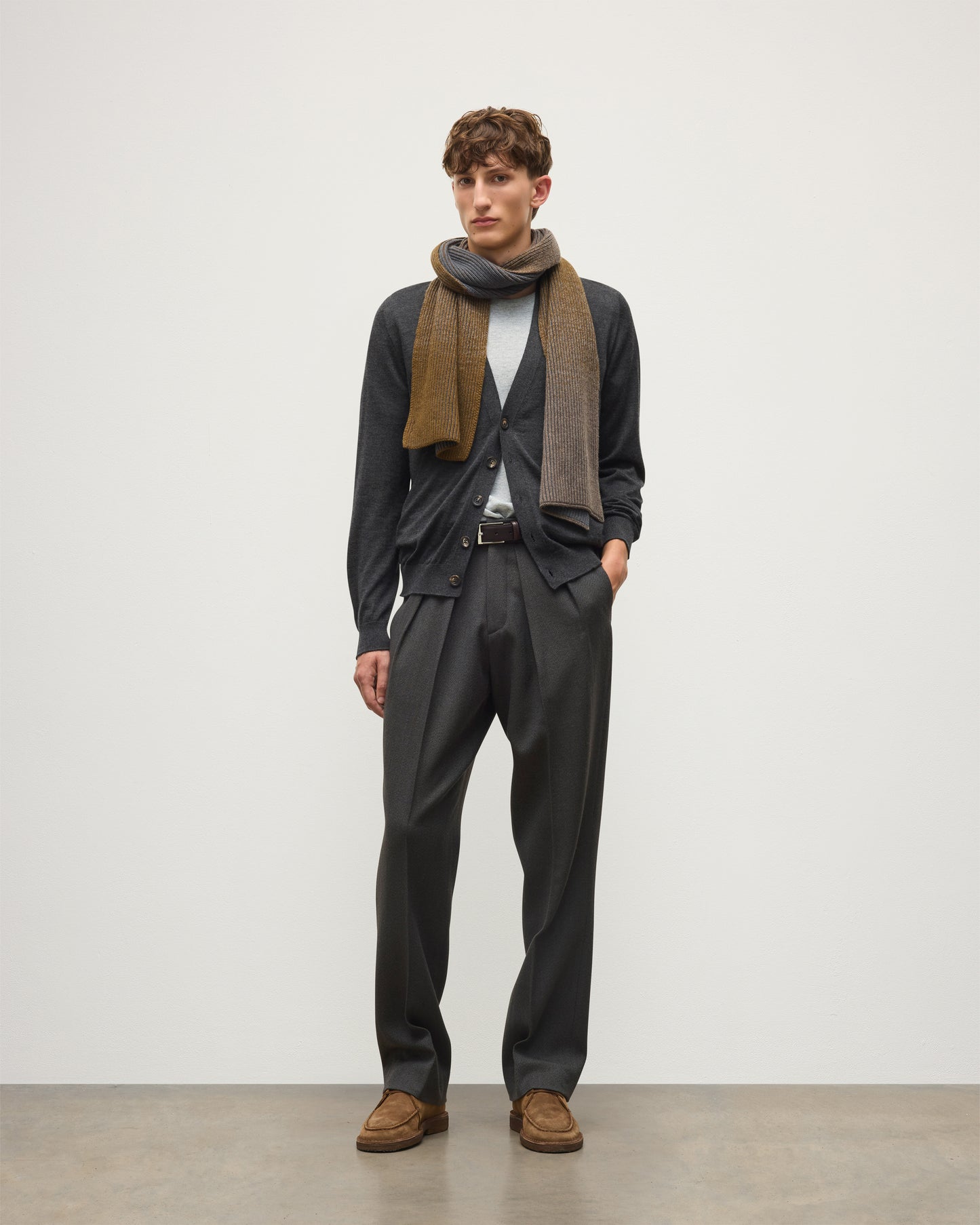 A man modeling a dark charcoal gray suit, featuring a dark gray cardigan, matching trousers, a light gray t-shirt, a brown and gray patterned scarf, and brown suede shoes, standing against a plain light gray studio background.  JOHNSTONS OF ELGIN CASHMERE RIBBED OMBRE PIONEER SCARF GREY 100% CASHMERE HAA03641Q26256ONE