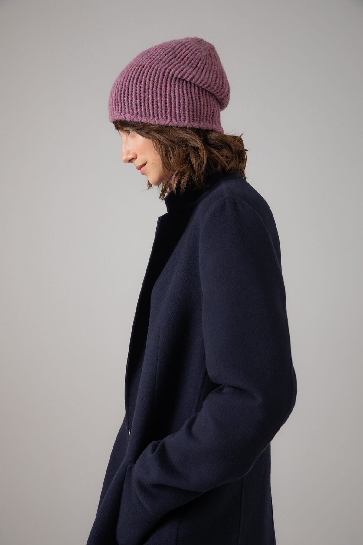 Johnstons of Elgin’s Heather pink Chunky Jersey Cashmere Hat on model wearing navy coat on a grey background HAB03196HE4307