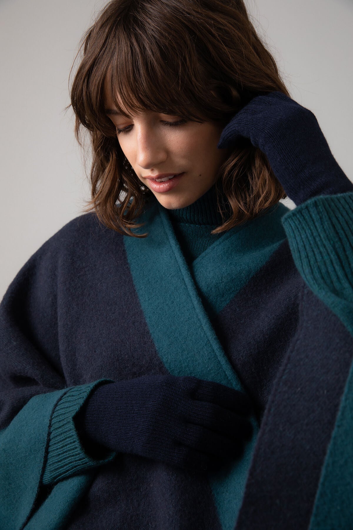 Model wearing Johnstons of Elgin Merino Wool Cape with Contrast Border in Mallard worn matching sweater and Navy Cashmere Gloves on a grey background TD000230RU7358ONE