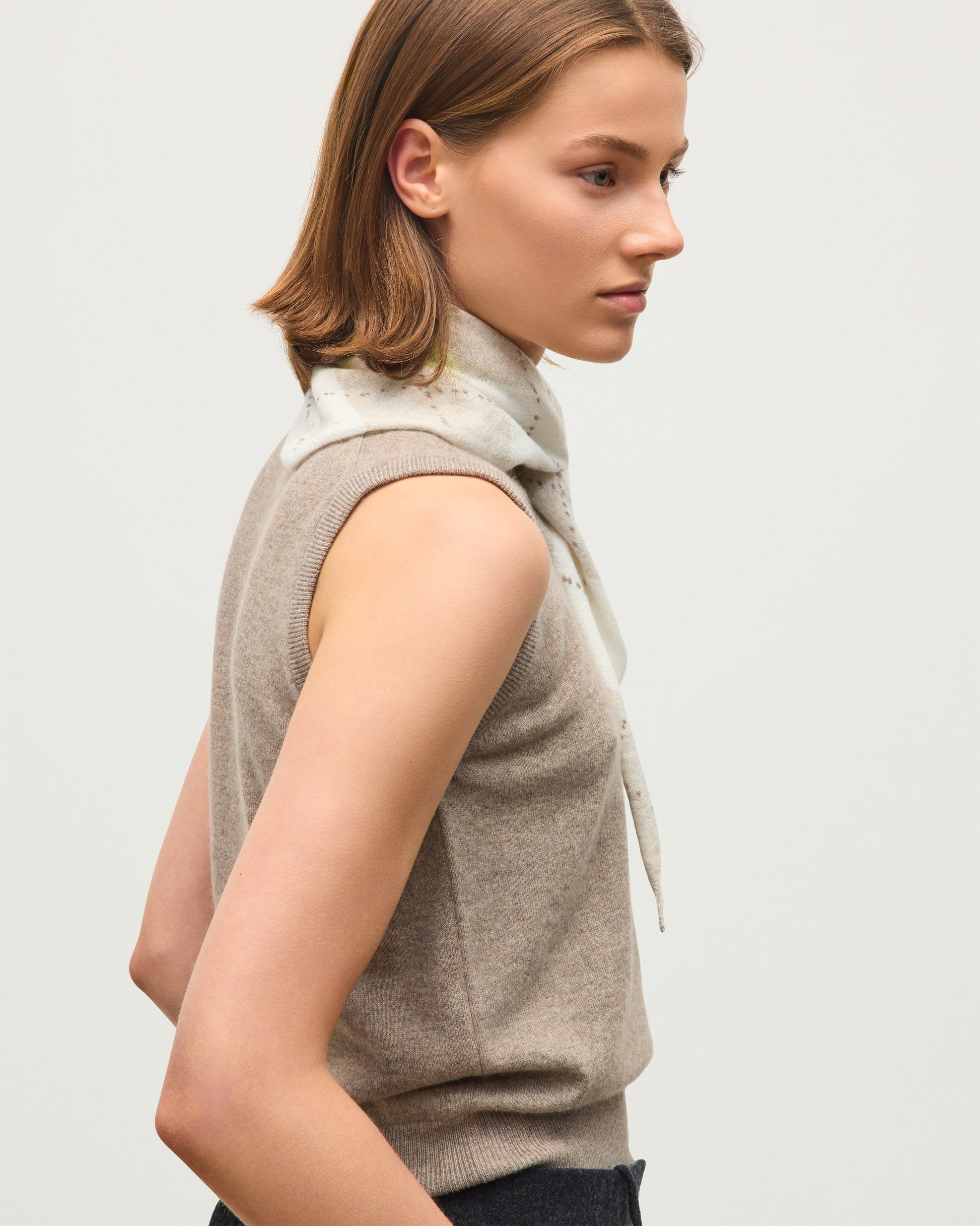 A woman shown in side profile wearing a sleeveless taupe knit vest over a light beige high-neck knit top, against a plain light gray studio background. JOHNSTONS OF ELGIN CASHMERE NECK TIE ARGYLE CLAN BANDANA NATURAL 100% CASHMERE HAP03612JU1629ONE
