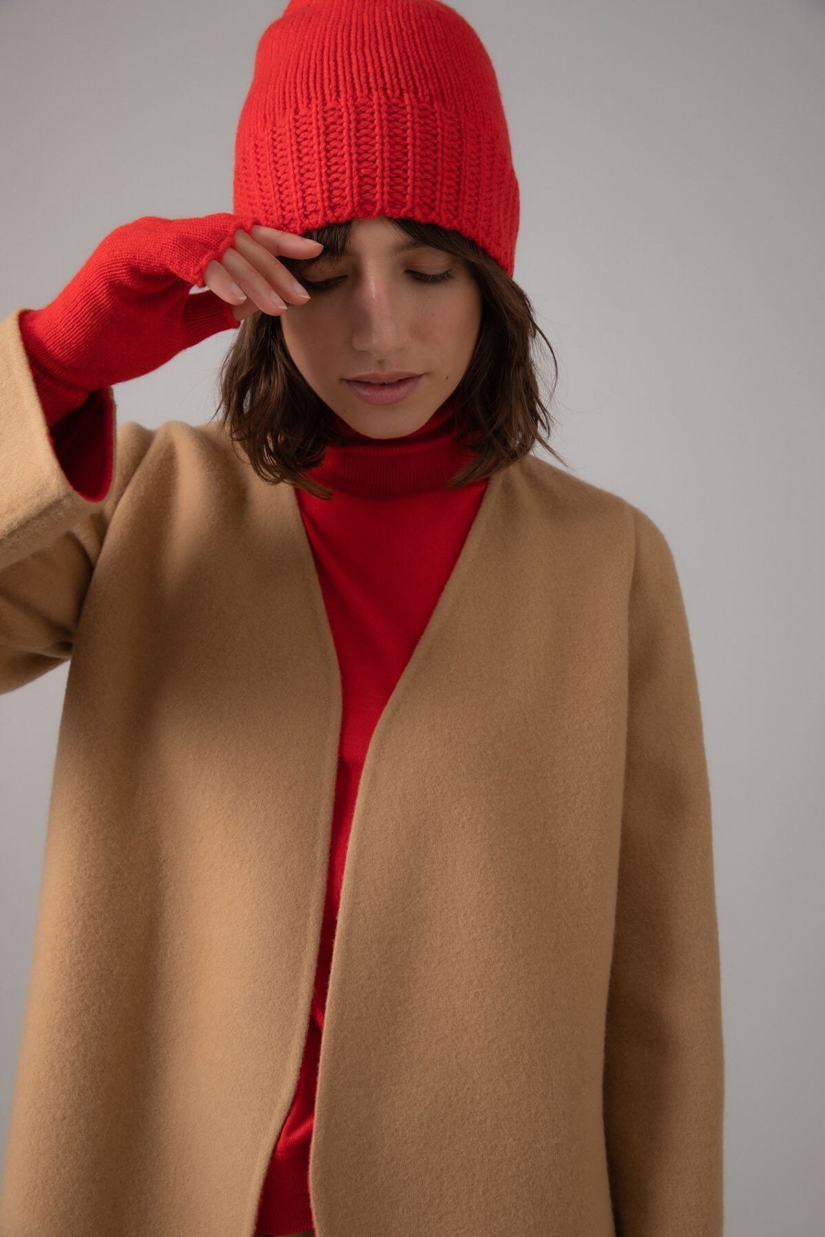 Johnstons of Elgin’s Orkney Red Women's Fingerless Cashmere Gloves on model wearing camel cashmere coat with red cashmere beanie HAY02223SE0661