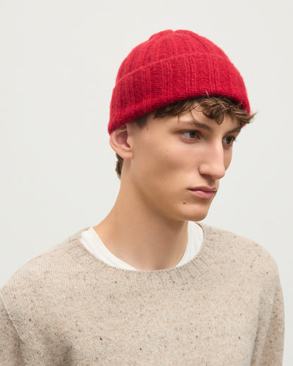 A young man wearing a bright red ribbed knit beanie and a light beige textured knit sweater, photographed against a clean, light gray studio background. JOHNSTONS OF ELGIN BRUSHED CASHMERE SILK FLAT RIB BHALTAIR BEANIE RED 74% CASHMERE, 26% SILK HBC01196HE4389ONE