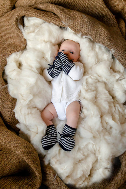 Baby wearing Johnstons of Elgin Hand Knitted Cashmere Baby Booties with crotched trims in Navy & White on a background of Cashmere fibres in a bale 746330333