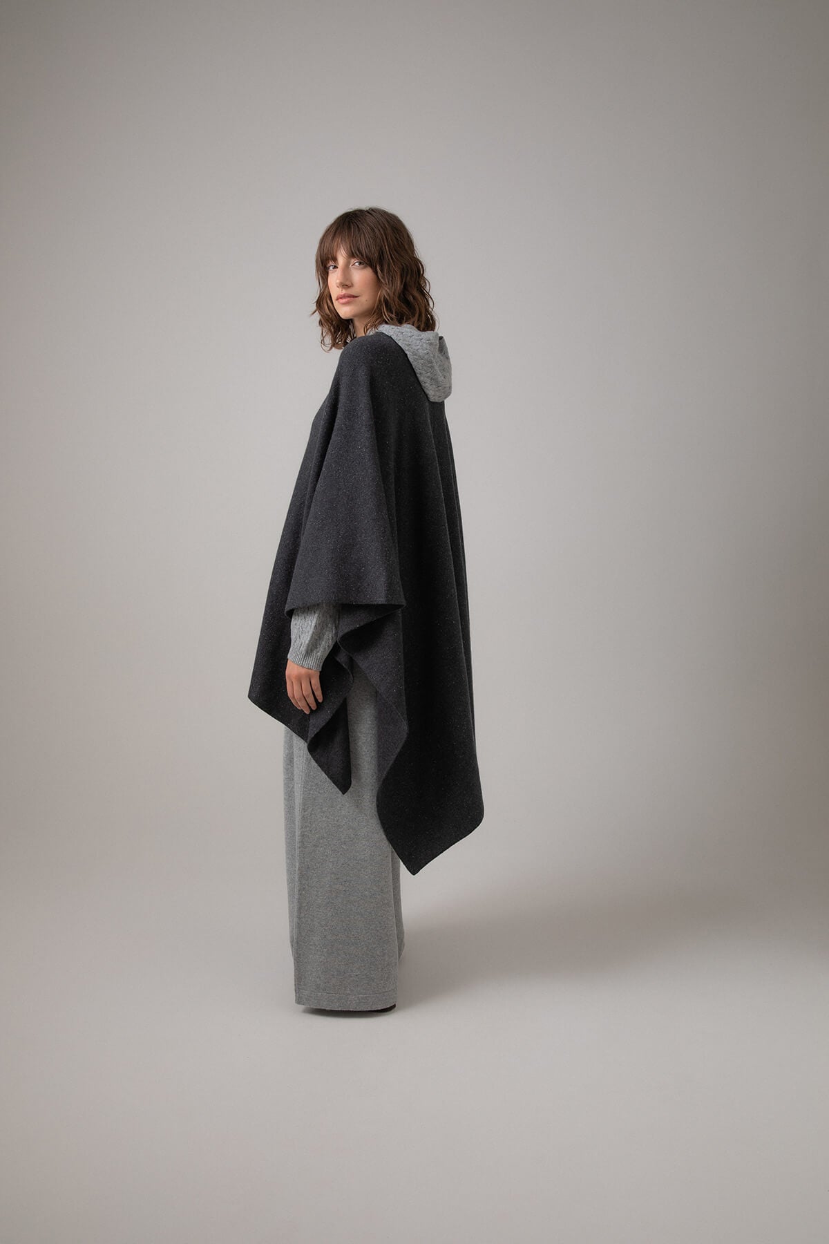 Side view of Johnstons of Elgin Double Face Donegal Cashmere Cape in Charcoal worn with Grey Cable Cashmere Hoodie and Wide Leg Joggers on a grey background KAA05102Q23681ONE