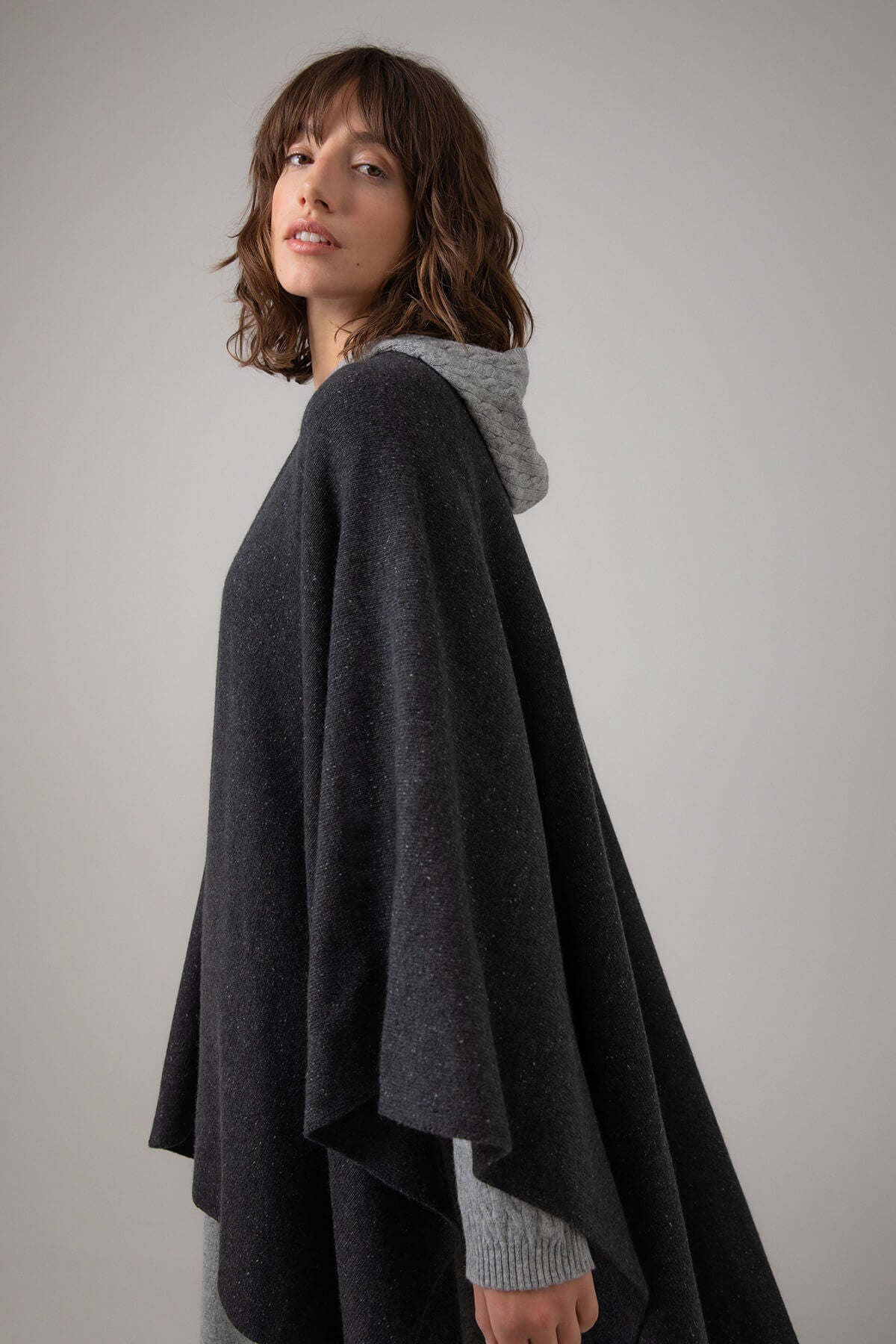 Side of a Johnstons of Elgin Double Face Donegal Cashmere Cape in Charcoal worn with Grey Cable Cashmere Hoodie on a grey background KAA05102Q23681ONE