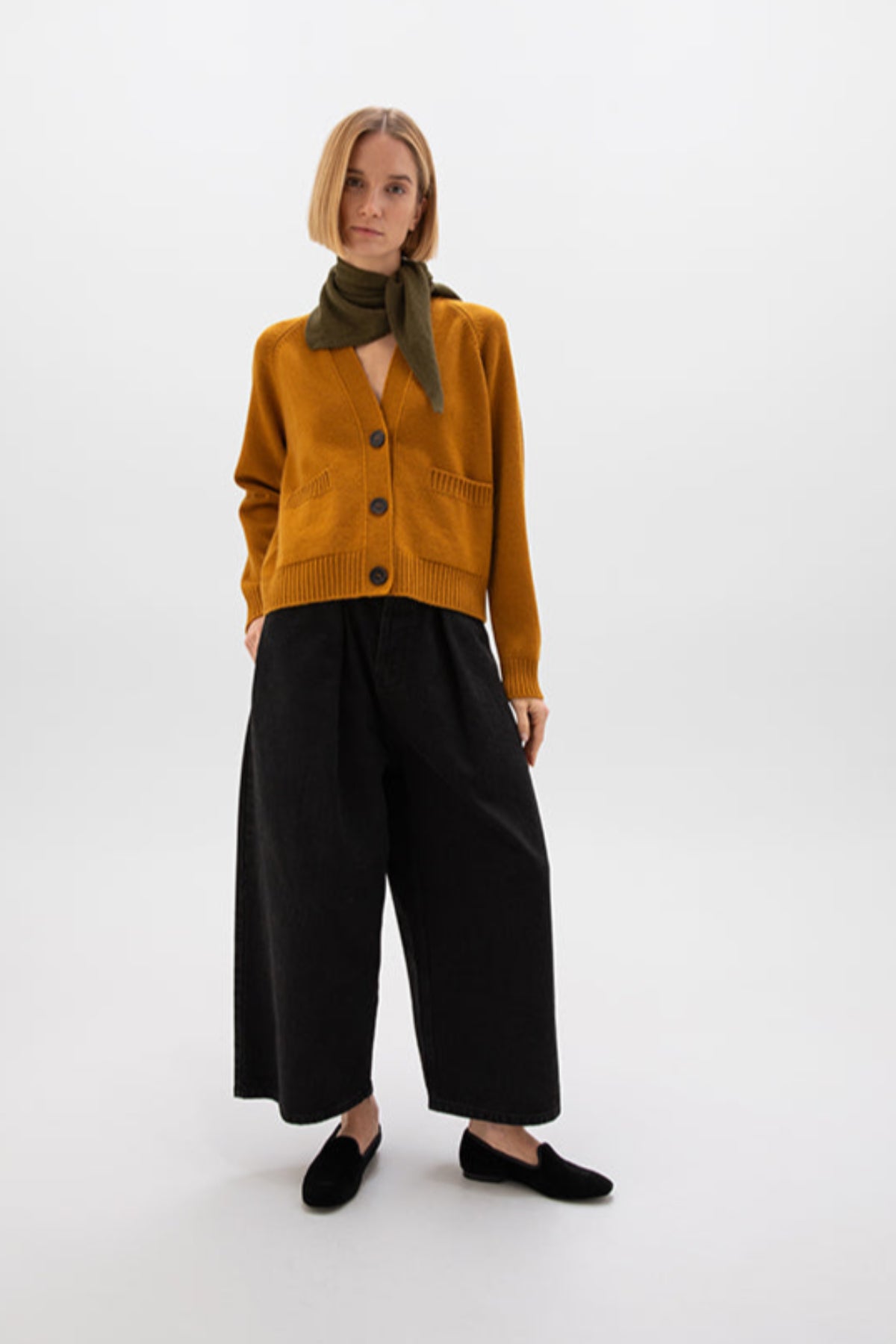 Person wearing a mustard-coloured cardigan, black pants, and a scarf on a white background KAA05320SF4347