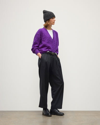 A model stands in profile wearing a purple cable-knit V-neck sweater, black wide-leg trousers, black leather shoes, a black beanie, and a thin black belt with a gold buckle, set against a minimalist off-white studio background. JOHNSTONS OF ELGIN KYLESKU DEEP VEE SWEATER CASHMERE CABLE DEEP VEE SWEATER PURPLE 100% CASHMERE KAA05728SD5372