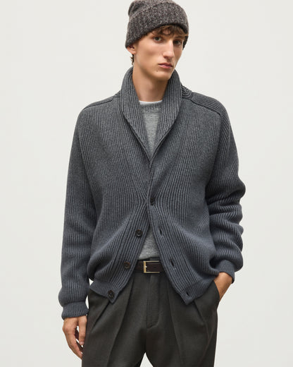 A man wearing a dark gray ribbed knit cardigan with a shawl collar, a gray beanie, a light gray t-shirt, and dark gray trousers, photographed against a light gray studio background. JOHNSTONS OF ELGIN PIONEER SHAWL CARDIGAN CASHMERE RIBBED OMBRE SHAWL COLLAR CARDIGAN - 6 PLY GREY 100% CASHMERE KAA05750Q26318