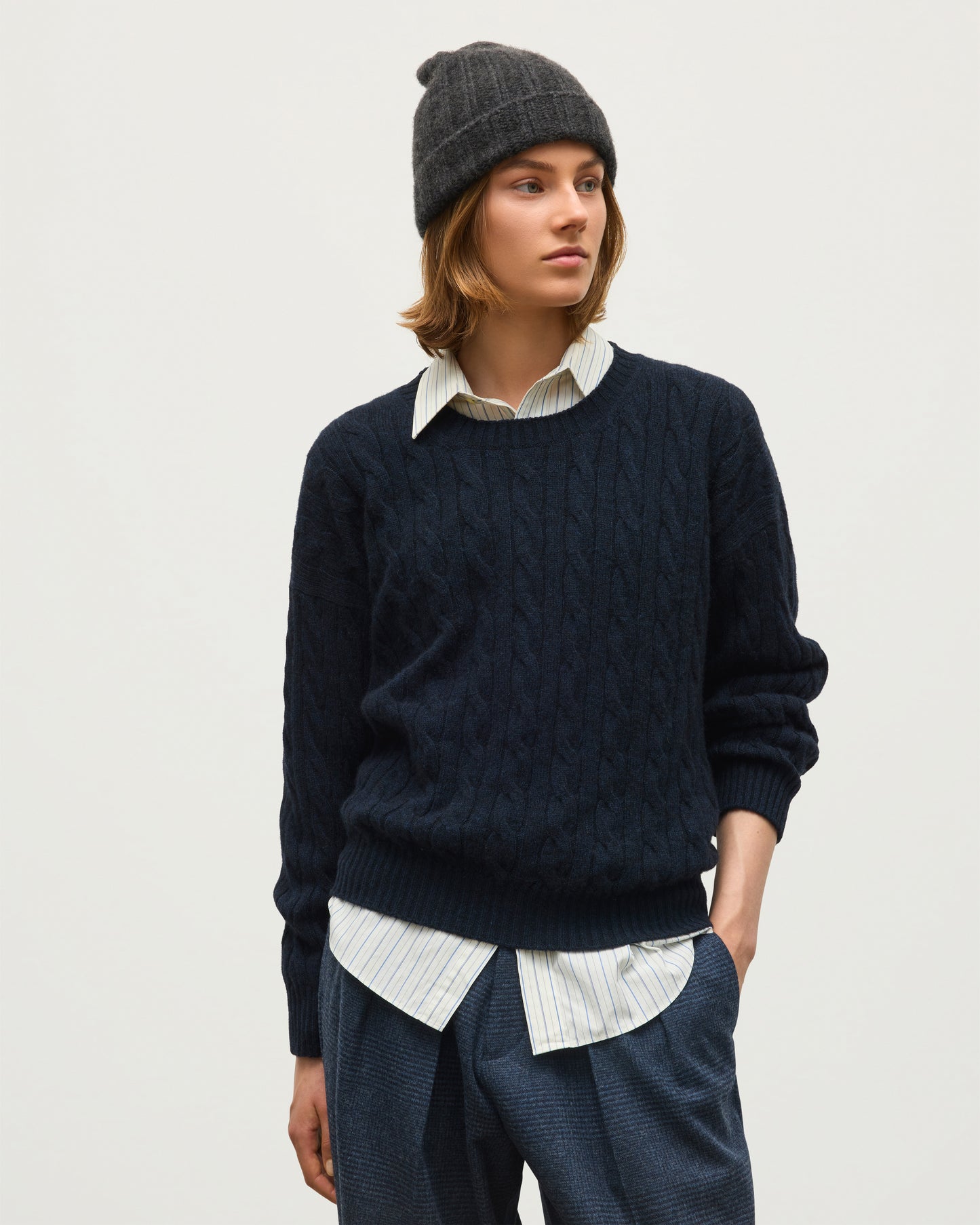 A model wearing a dark navy cable-knit crewneck sweater over a striped collared shirt, paired with dark trousers and a dark knit beanie, against a plain light gray background. JOHNSTONS OF ELGIN KYLESKU CREW SWEATER CASHMERE CABLE ROUND NECK SWEATER DARK BLUE 100% CASHMERE KAA06028HD7347