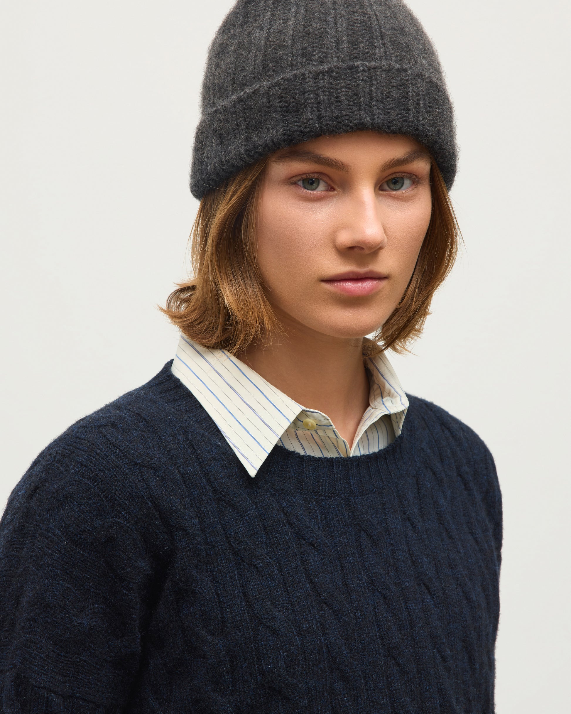 A close-up of a model wearing a dark navy cable-knit sweater over a striped button-down shirt, paired with a dark gray knit beanie, against a plain light background. JOHNSTONS OF ELGIN KYLESKU CREW SWEATER CASHMERE CABLE ROUND NECK SWEATER DARK BLUE 100% CASHMERE KAA06028HD7347