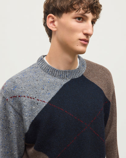 A close-up of a man wearing a color-blocked knit sweater with a speckled gray shoulder, a dark navy center panel, and a brown sleeve, accented by red dashed lines. JOHNSTONS OF ELGIN MONTEITH CREW SWEATER DONEGAL CASHMERE EXPLODED ARGYLE CREW SWEATER GREY 100% CASHMERE KAA06033JU1639