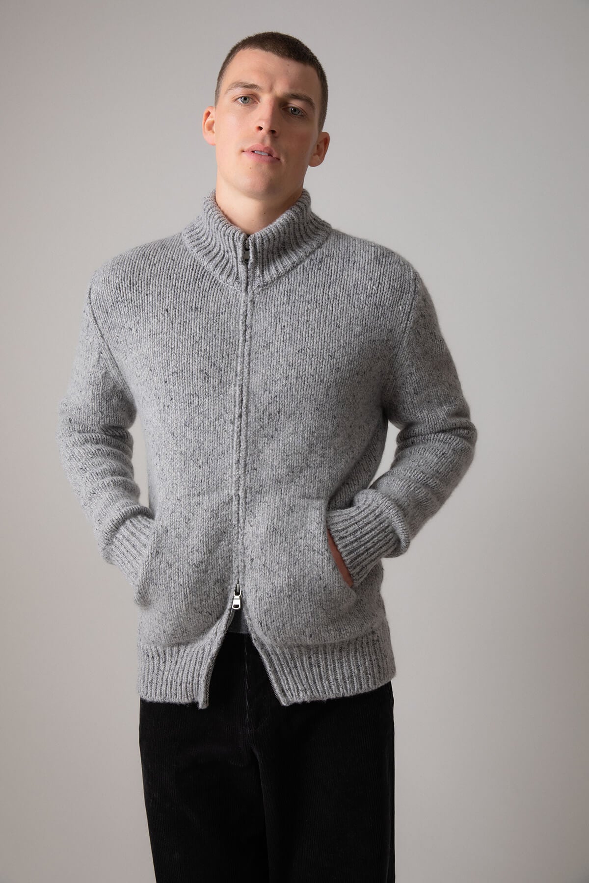 Johnstons of Elgin’s Men's Cashmere Donegal Zip Turtle Neck Cardigan in Light Grey on model wearing black trousers on a grey background KAB05115Q23711
