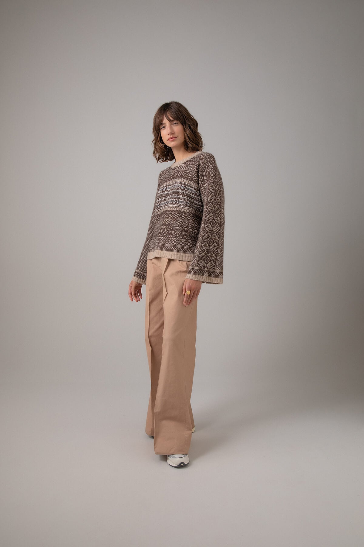 Johnstons of Elgin Women's Round Neck Traditional Sanquhar Fairisle Cashmere Sweater in Oatmeal worn with Camel Trousers on a grey background KAC05099Q23724