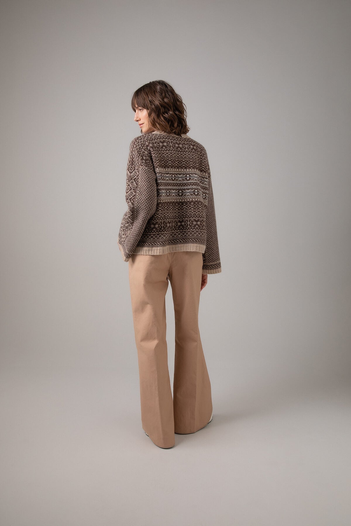 Back of Johnstons of Elgin Women's Round Neck Traditional Sanquhar Fairisle Cashmere Sweater in Oatmeal worn with Camel Trousers on a grey background KAC05099Q23724