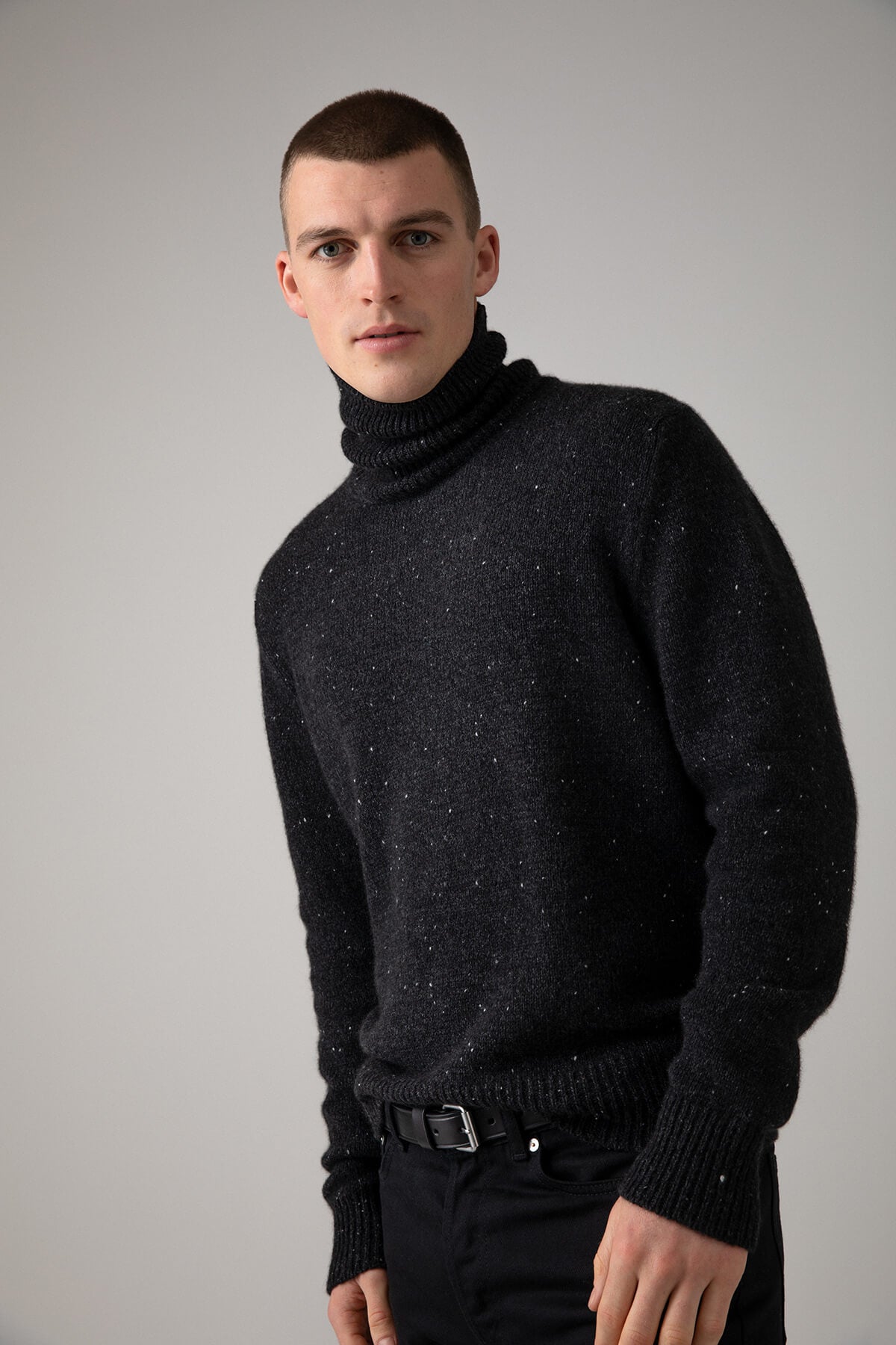 Johnstons of Elgin’s Men's Cashmere Donegal Roll Neck Jumper in Charcoal grey on model wearing black trousers on a grey background KAC05111002675