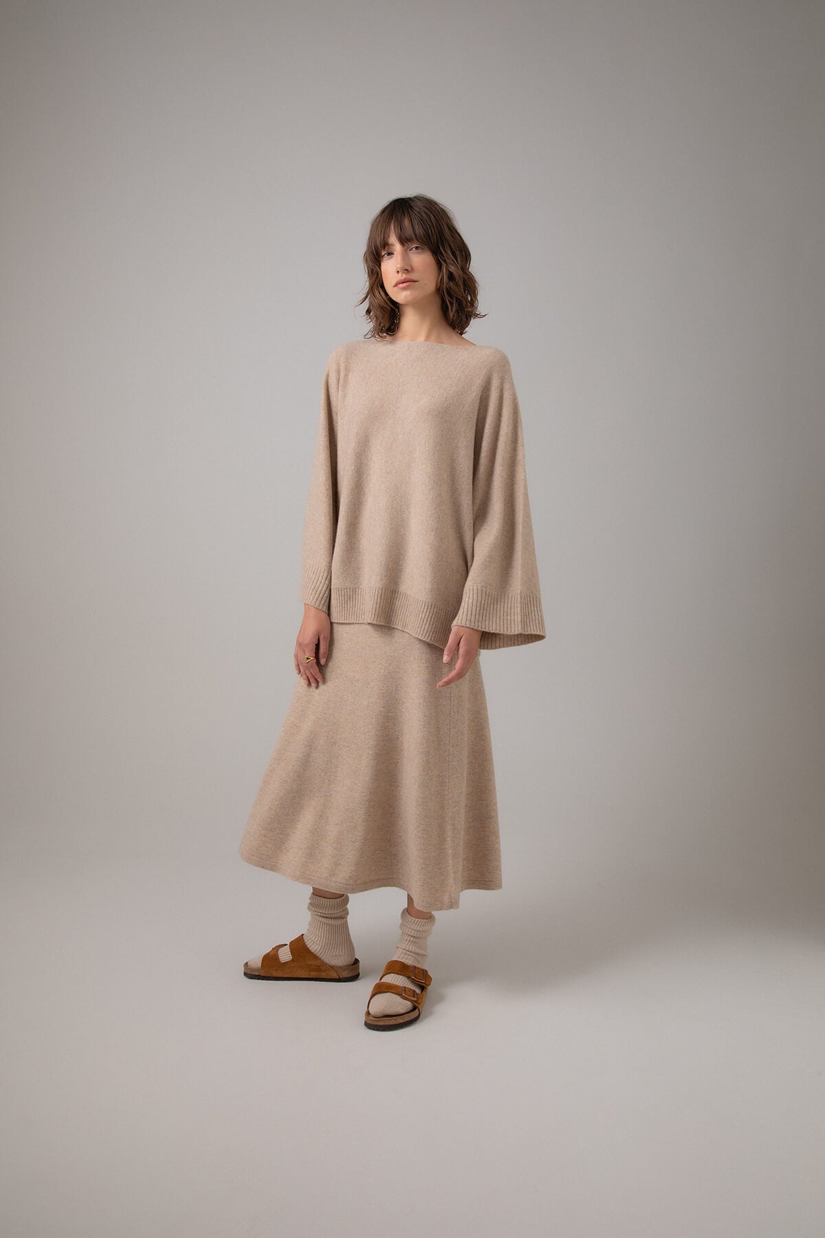 Johnstons of Elgin Boat Neck Cashmere Cape Jumper in Oatmeal worn with a matching A-Line Skirt on a grey background KAI05048HB0210
