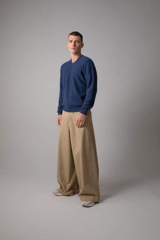 Johnstons of Elgin’s Men’s Textured Cashmere High V Neck Jumper in ocean blue on model wearing wide camel trousers on a grey background KAI05118HD7244