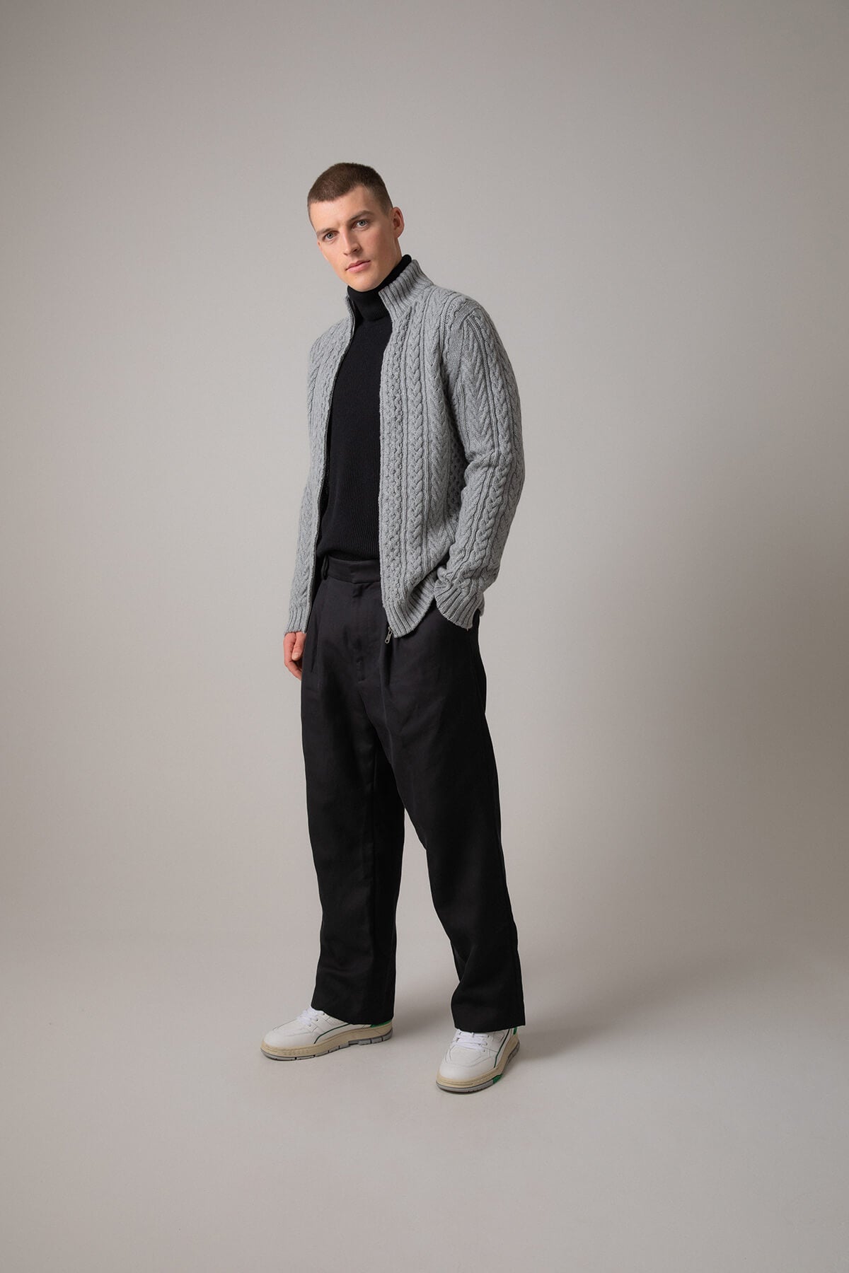 Johnstons of Elgin’s Men's Aran Cable Cashmere Zip Cardigan in Light Grey on model wearing black trousers on a grey background KAI05119Q23698
