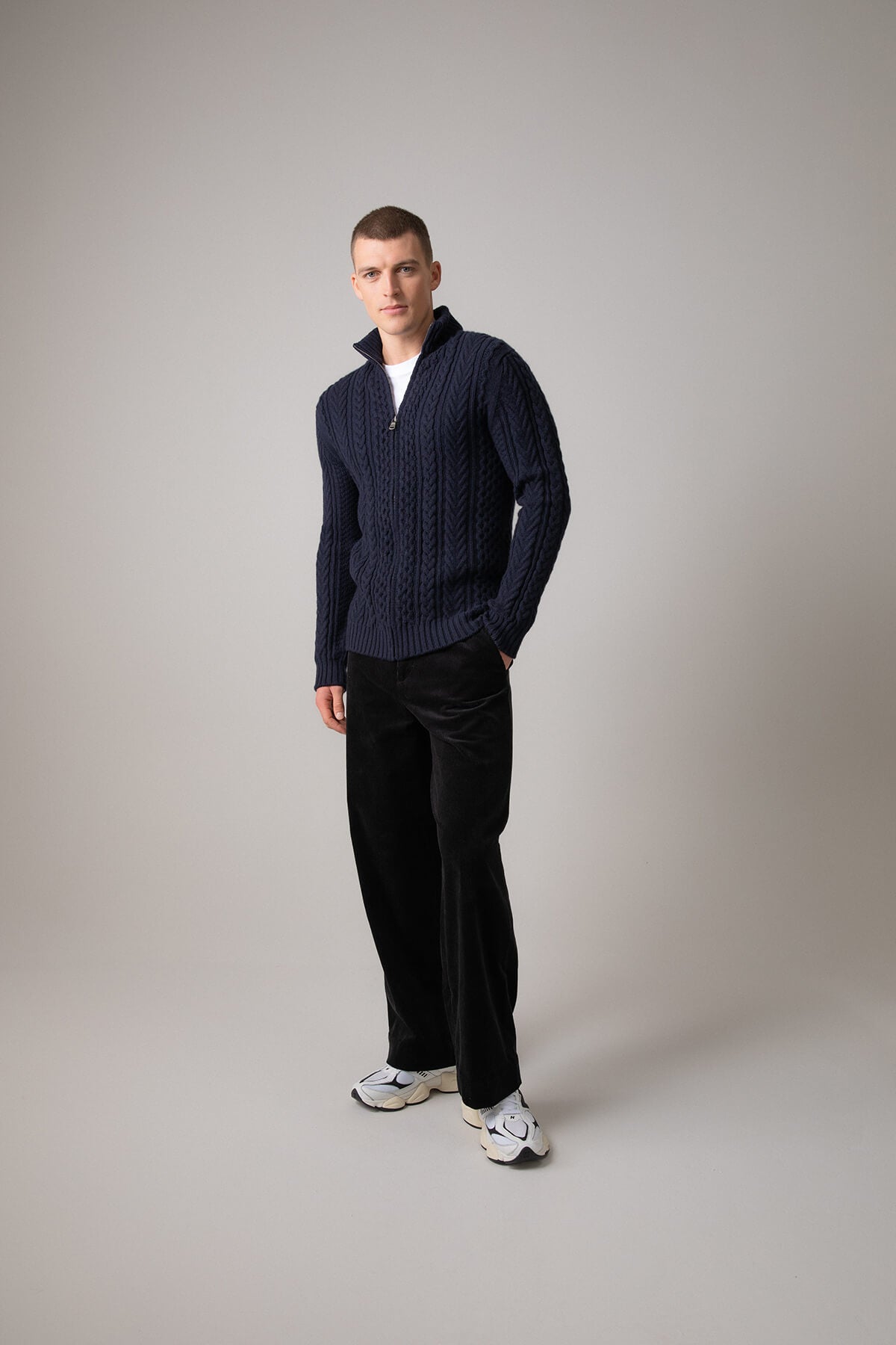 Johnstons of Elgin’s Men's Aran Cable Cashmere Zip Cardigan in Dark Navy on model wearing black trousers on a grey background KAI05119Q23706