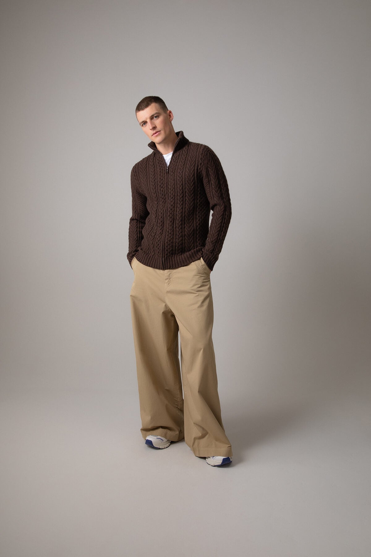 Johnstons of Elgin’s Men's Aran Cable Cashmere Zip Cardigan in Peat on model wearing wide camel trousers on a grey background KAI05119Q23708
