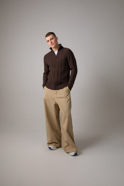 Johnstons of Elgin’s Men's Aran Cable Cashmere Zip Cardigan in Peat on model wearing wide camel trousers on a grey background KAI05119Q23708