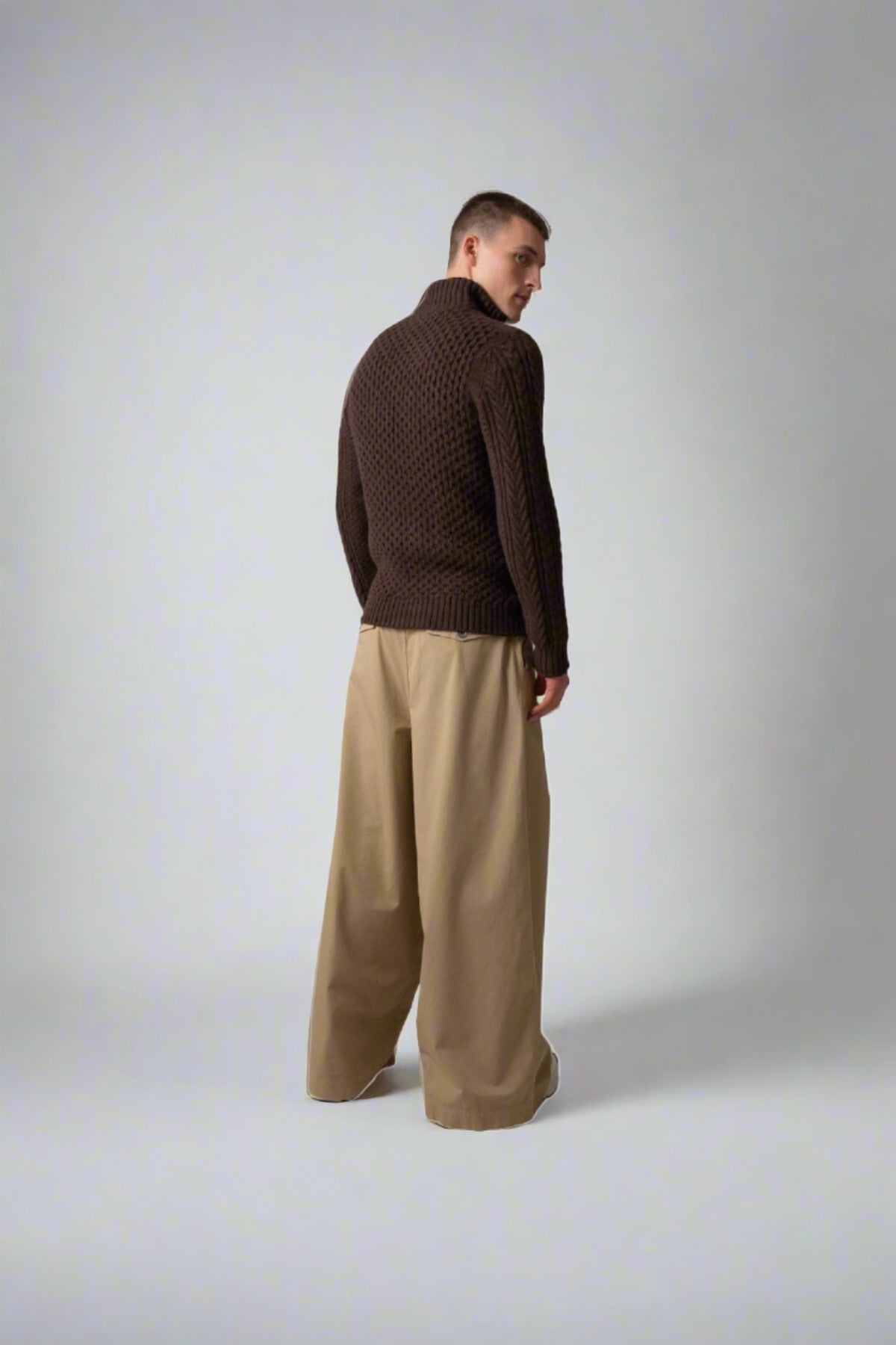 Johnstons of Elgin’s Men's Aran Cable Cashmere Zip Cardigan in Peat on model wearing wide camel trousers on a grey background KAI05119Q23708