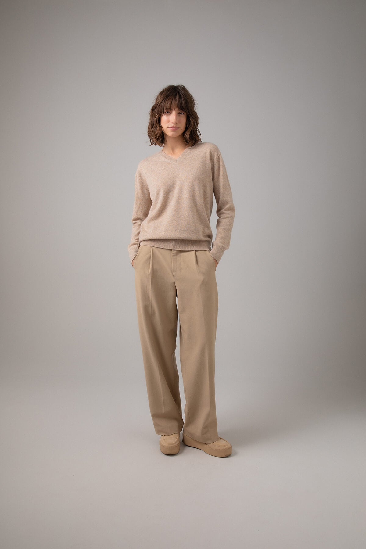 Johnstons of Elgin Women's V Neck Cashmere Jumper in Oatmeal worn with Camel Trousers on a grey background KAI05140HB0210