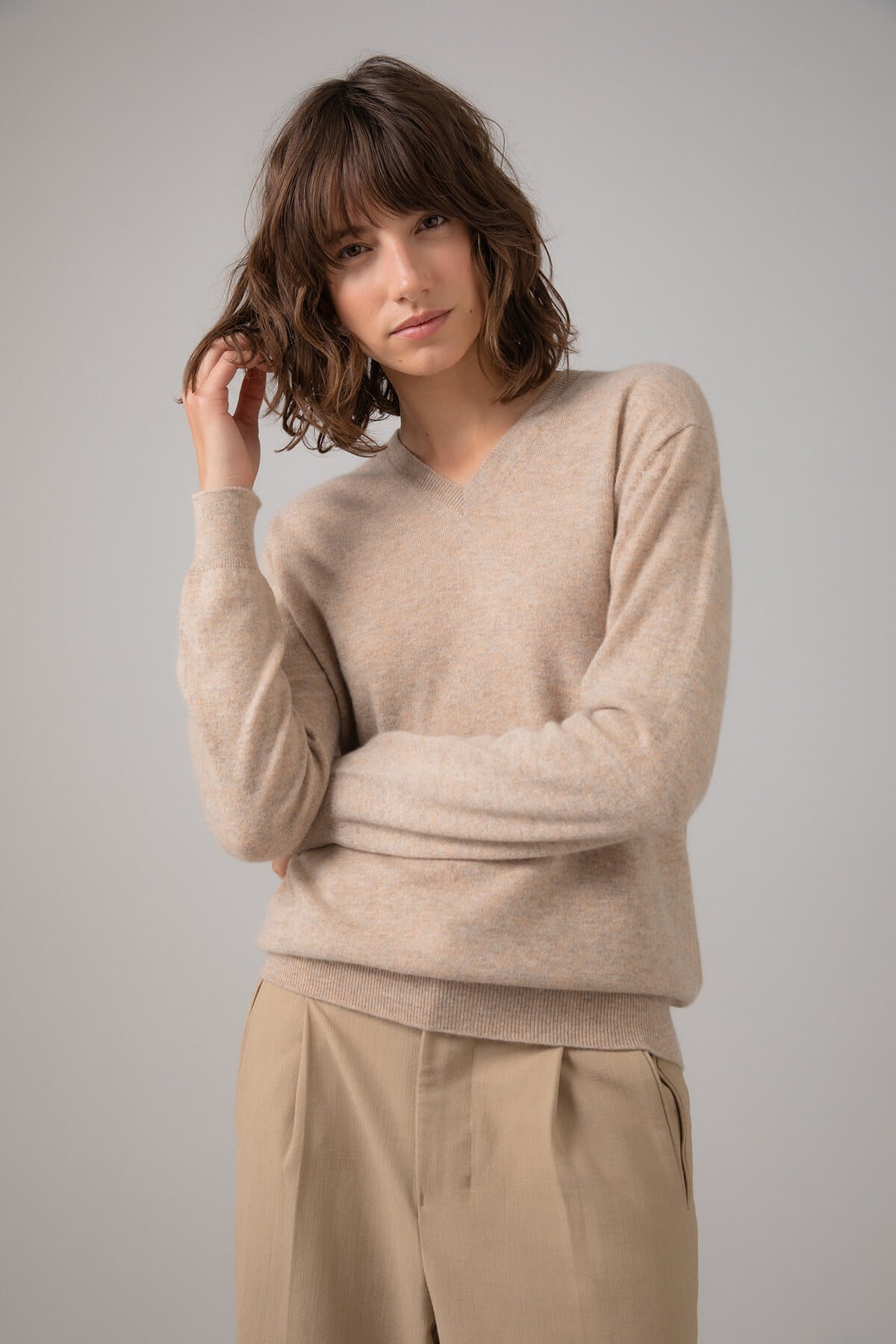 Model wearing Johnstons of Elgin Women's V Neck Cashmere Jumper in Oatmeal worn with Camel Trousers on a grey background KAI05140HB0210