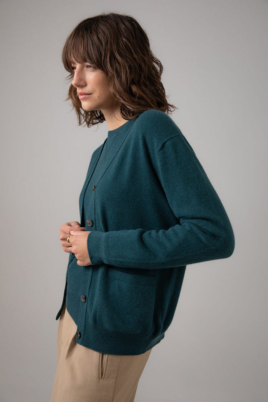 Side view of a Johnstons of Elgin Women's Cashmere Boyfriend Cardigan in Mallard green worn with a matching Crew Neck on a grey background KAI05141HC7126