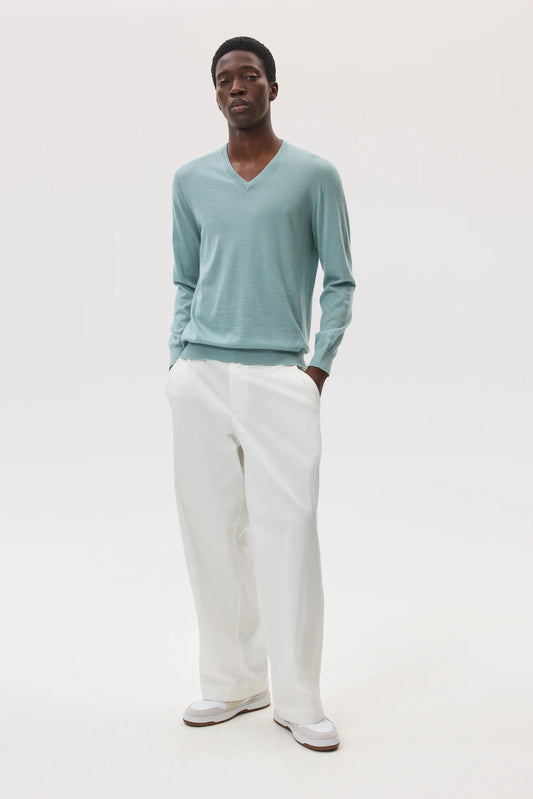 Male model wearing Johnstons of Elgin SS25 Unisex Knitwear Sage Superfine Cashmere V Neck Sweater with white trousers on a studio backdrop of white KAI05242SC0249
