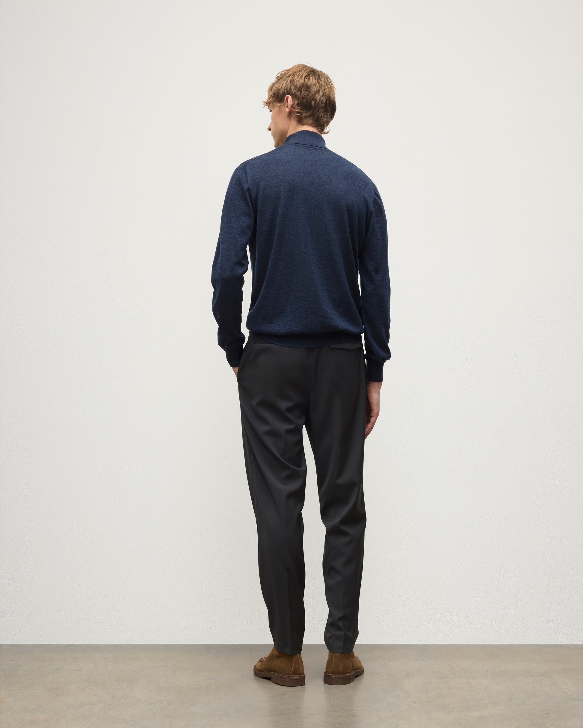 Person wearing a navy sweater and black pants standing against a white wall. KAI05333HD7350