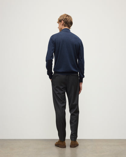 Person wearing a navy sweater and black pants standing against a white wall. KAI05333HD7350