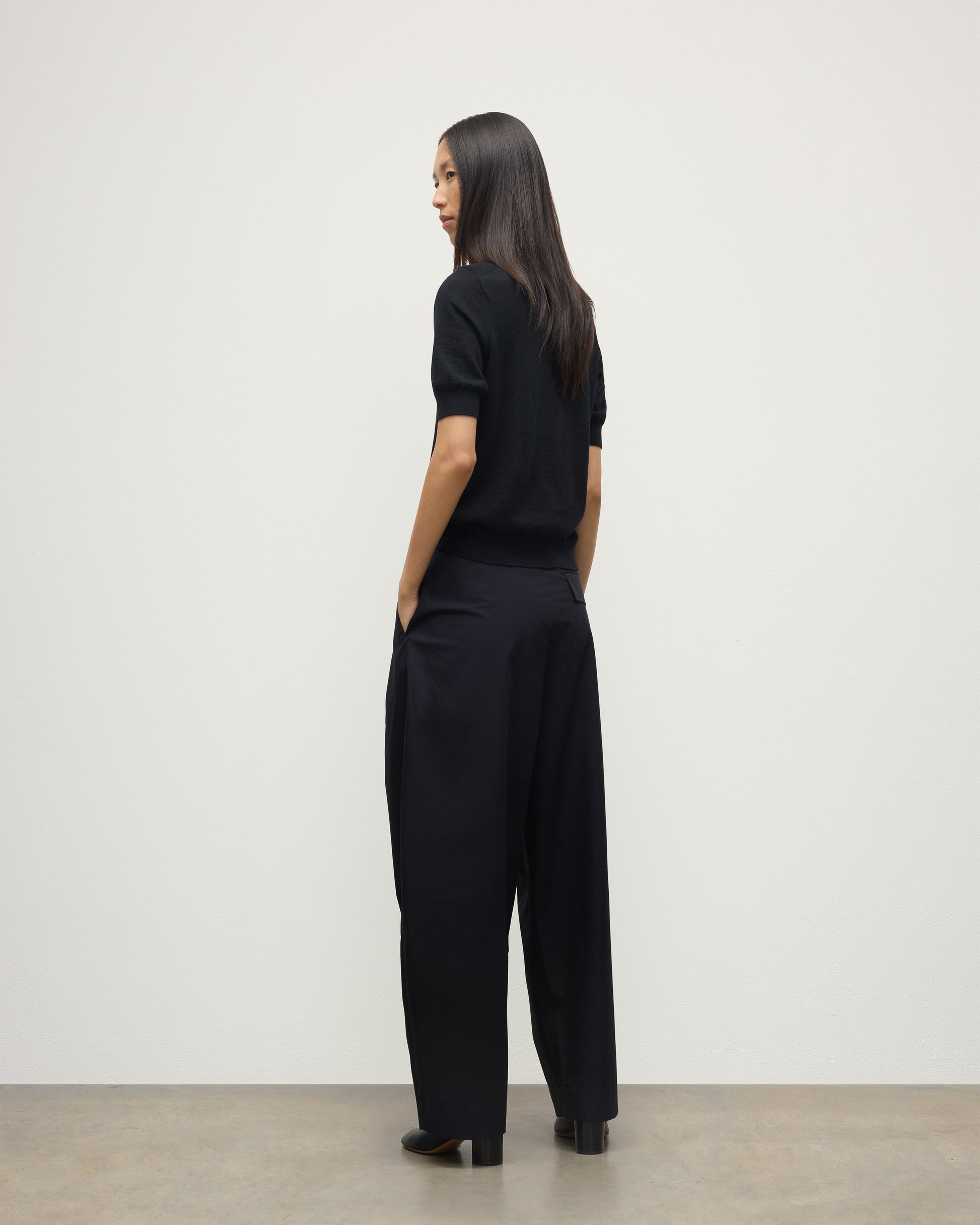 BACK OF A MODEL WEARS JOHNSTONS OF ELGIN WOMENS HONOR SHORT SLEEVE CREW BLACK 100% SUPERFINE CASHMERE WITH BLACK TROUSERS AND BOOTS KAI05712SA7146