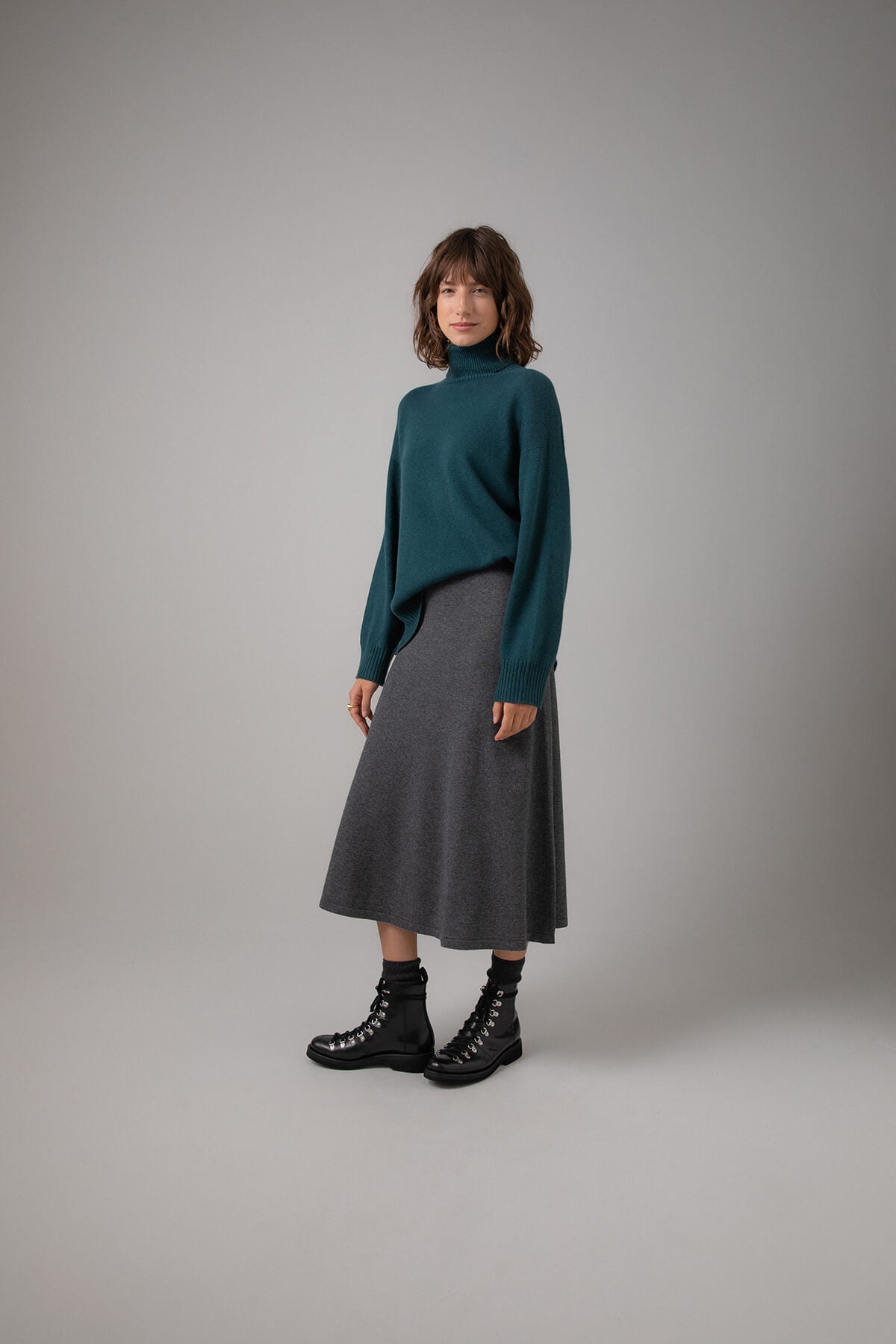 Side view of a Johnstons of Elgin Women's A-Line Cashmere Skirt in Mid Grey worn with a Mallard Roll Neck Sweater on a grey background KAP05097HA4181