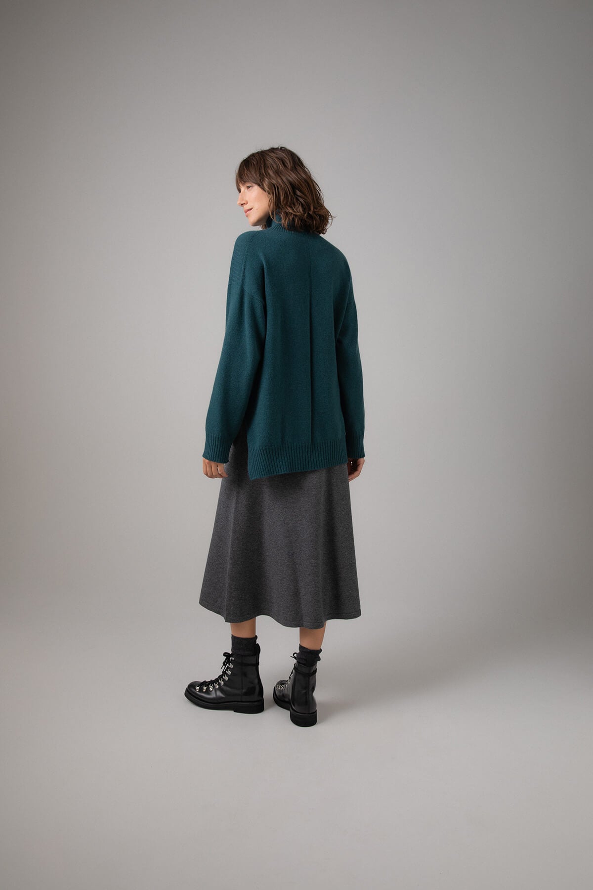 Back view of a Johnstons of Elgin Women's A-Line Cashmere Skirt in Mid Grey worn with a Mallard Roll Neck Sweater on a grey background KAP05097HA4181