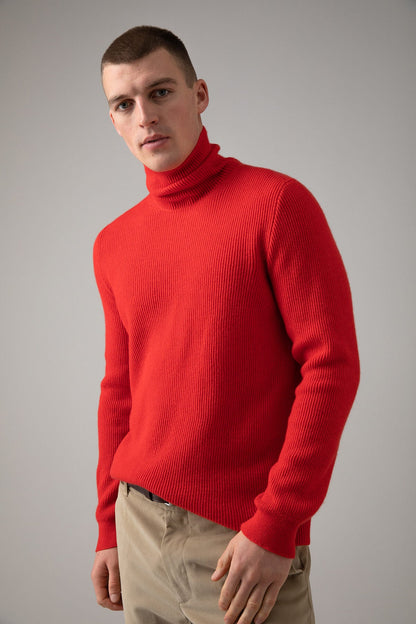 Johnstons of Elgin Men’s Ribbed Cashmere Roll Neck Jumper in Orkney Red on model wearing camel trousers on grey background KAP05107SE0661