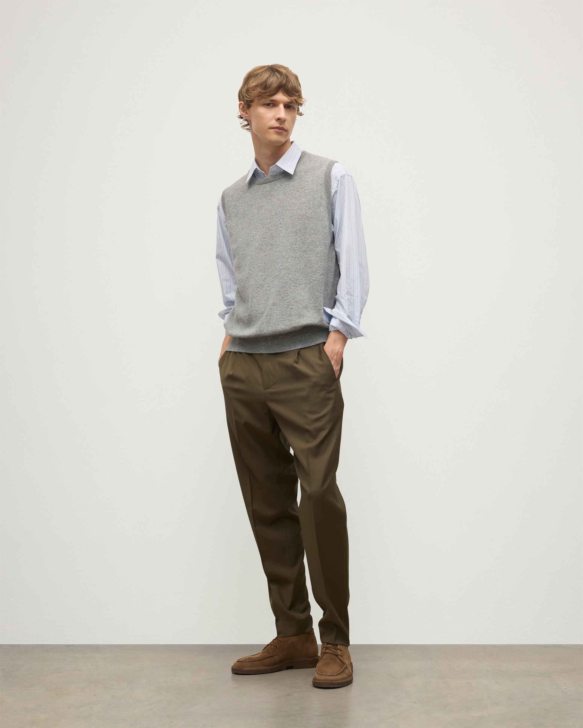 MODEL WEARS JOHNSTONS OF ELGIN MENS LENNOX SLEEVELESS CREW GREY 100% CASHMERE OVER A LIGHT BLUE SHIRT WITH OLIVE GREEN TROUSERS KAP05243HA0400