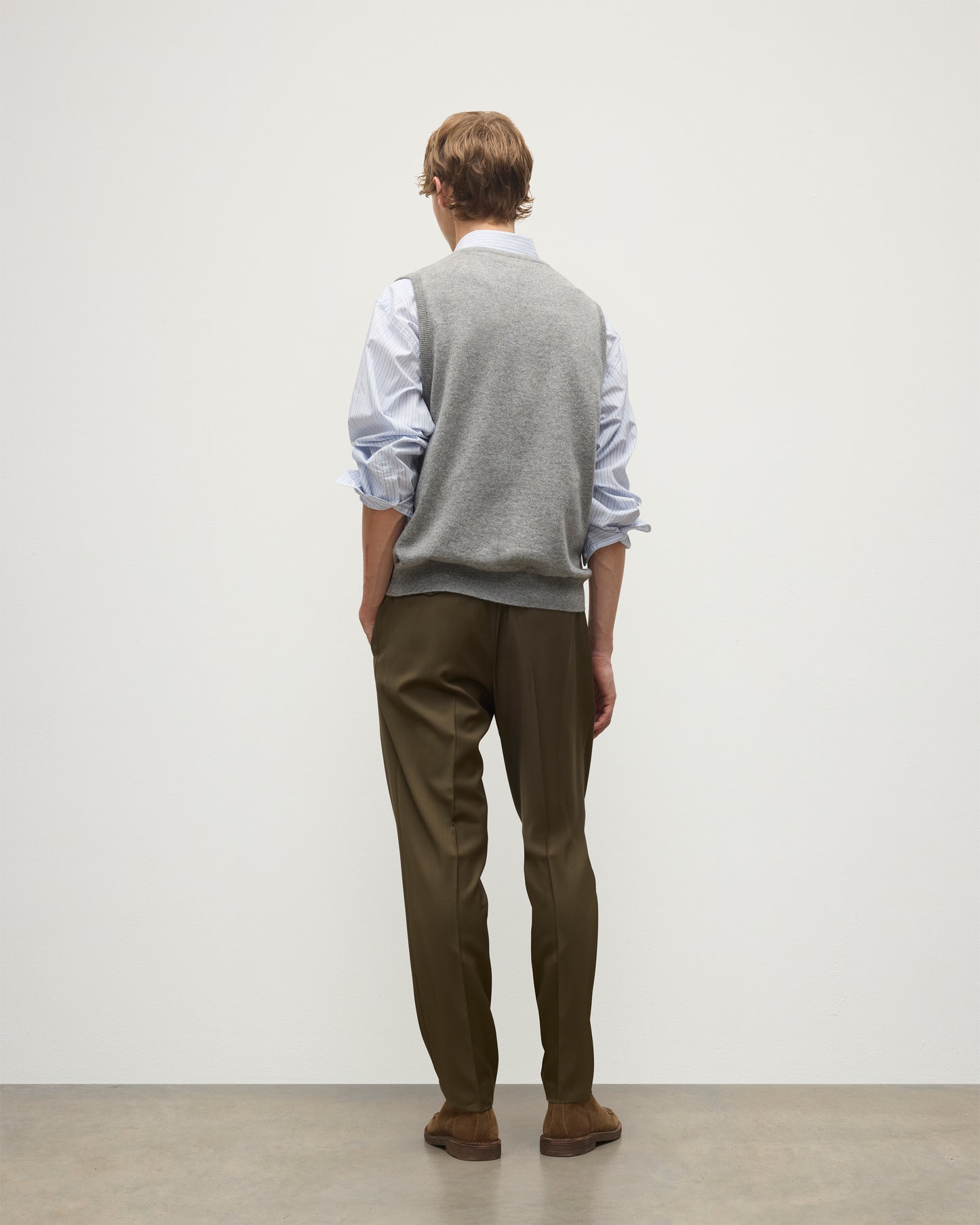 MODEL WEARS JOHNSTONS OF ELGIN MENS LENNOX SLEEVELESS CREW GREY 100% CASHMERE OVER A LIGHT BLUE SHIRT WITH OLIVE GREEN TROUSERS KAP05243HA0400