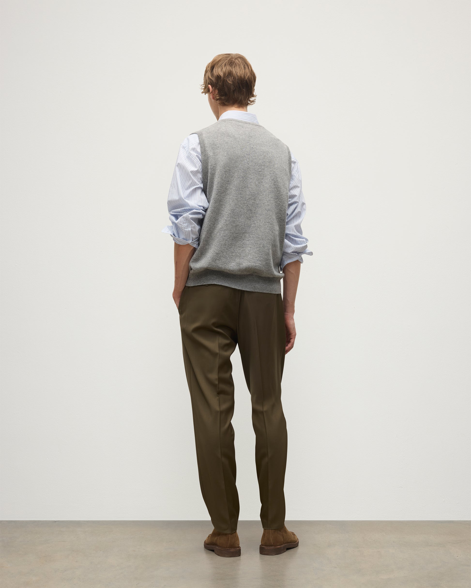 MODEL WEARS JOHNSTONS OF ELGIN MENS LENNOX SLEEVELESS CREW GREY 100% CASHMERE OVER A LIGHT BLUE SHIRT WITH OLIVE GREEN TROUSERS KAP05243HA0400