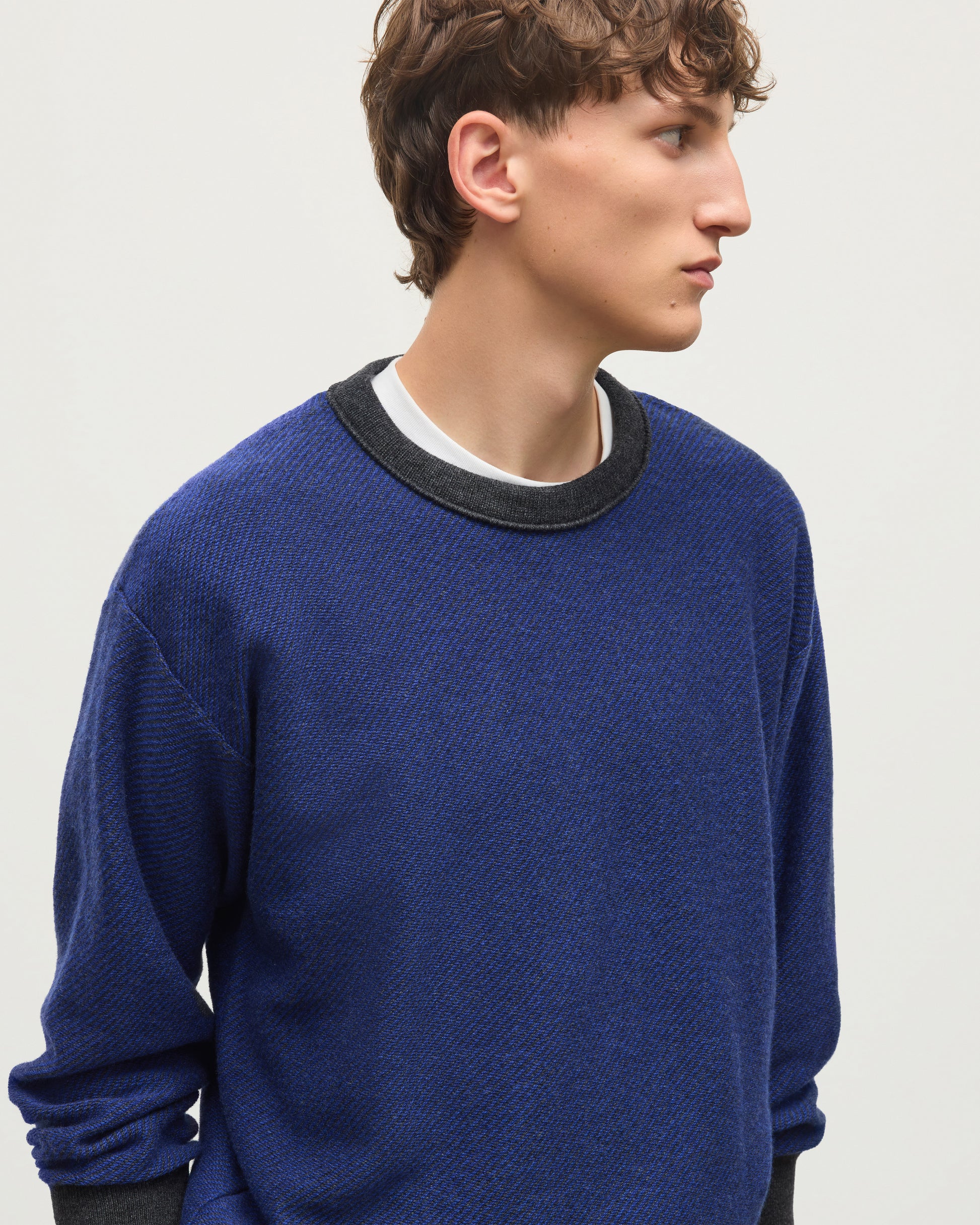 A man wearing a textured royal blue crewneck sweater with a contrasting black ribbed collar, layered over a white t-shirt, photographed against a light gray studio background. JOHNSTONS OF ELGIN LIDDELL SWEATSHIRT CASHMERE REVERSE TWILL STITCH SWEATSHIRT - 2 PLY GREY & BLUE 100% CASHMERE KAP05725Q26375