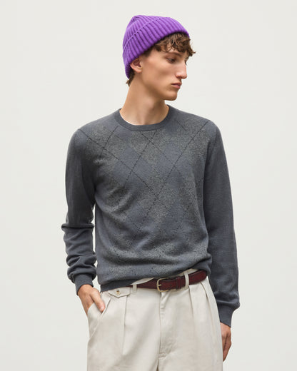A man wearing a dark gray argyle sweater, light beige trousers, a dark brown belt, and a purple knit beanie, standing against a plain light gray studio background. JOHNSTONS OF ELGIN ARGYLL CREW SWEATER CASHMERE ARGYLE INTARSIA CREW NECK SWEATER - 2 PLY GREY 100% CASHMERE KAP05747JU1636