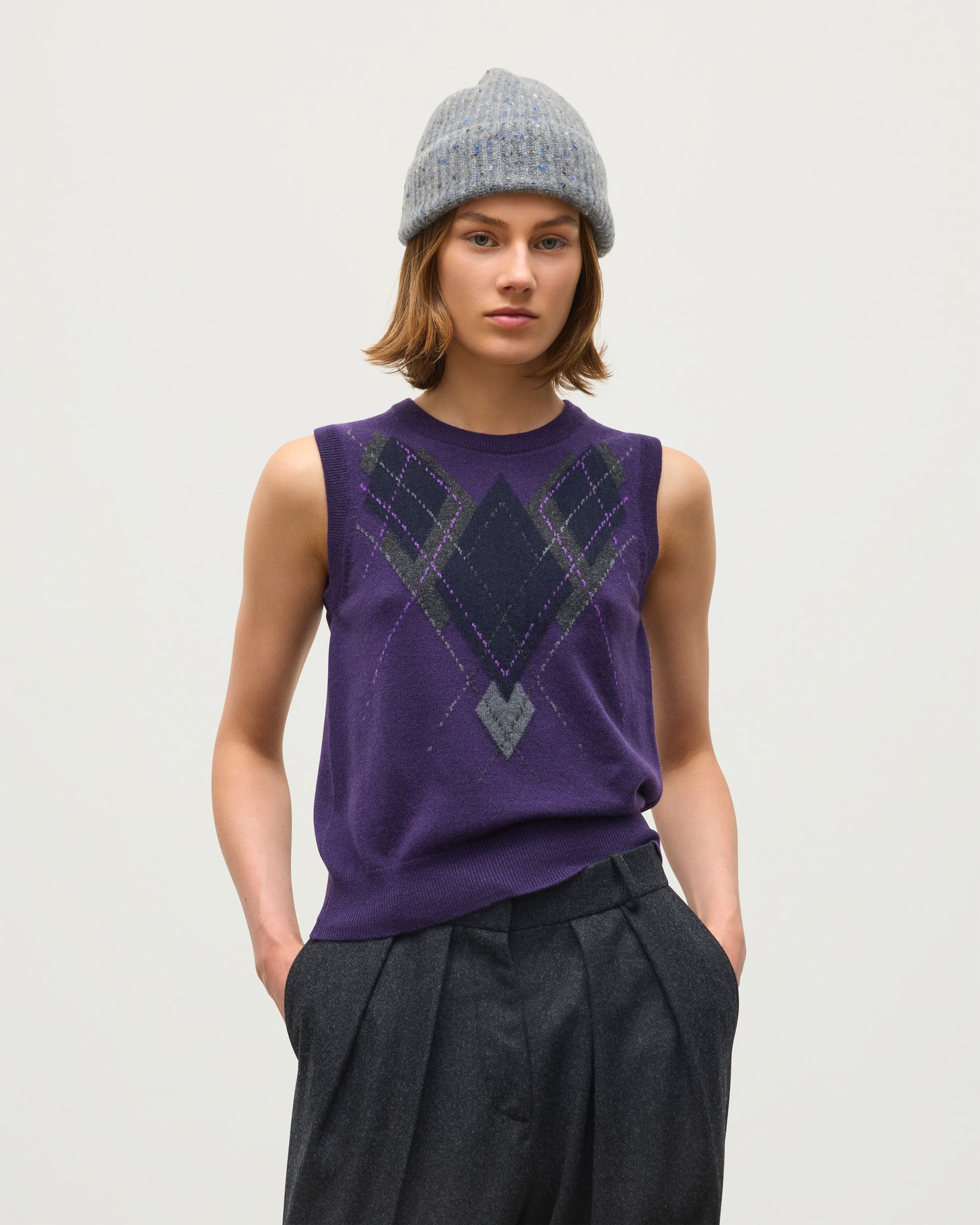 A model wearing a sleeveless purple knit top with a dark geometric argyle pattern on the chest, paired with dark pleated trousers and a gray knit beanie, against a plain light gray background. JOHNSTONS OF ELGIN LE JABOTS SLEEVELESS CREW PETITE CASHMERE ARGYLE INTARSIA ROUND NECK TANK - 2 PLY PURPLE 100% CASHMERE KAP05766JU1648