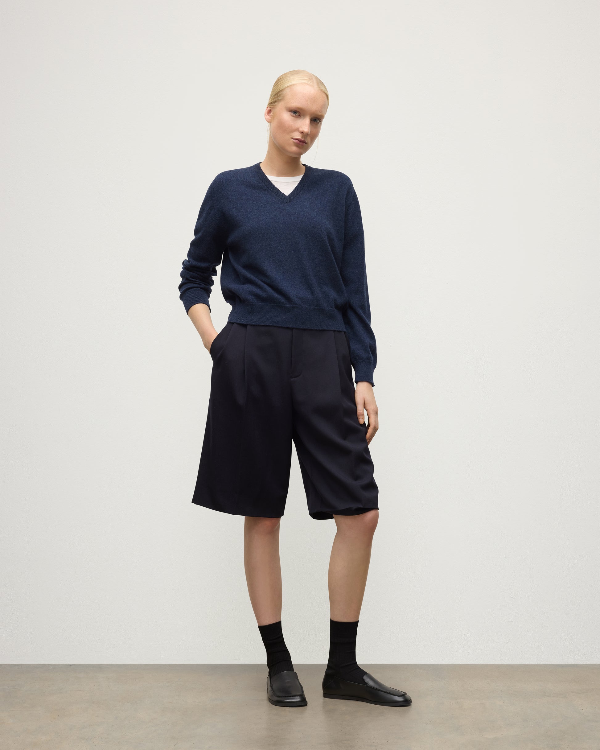 MODEL WEARING JOHNSTONS OF ELGIN WOMENS LUNAN CLASSIC VEE SWEATER NAVY 100% CASHMERE WITH LONG BLACK SHORTS KAP05807HD7350